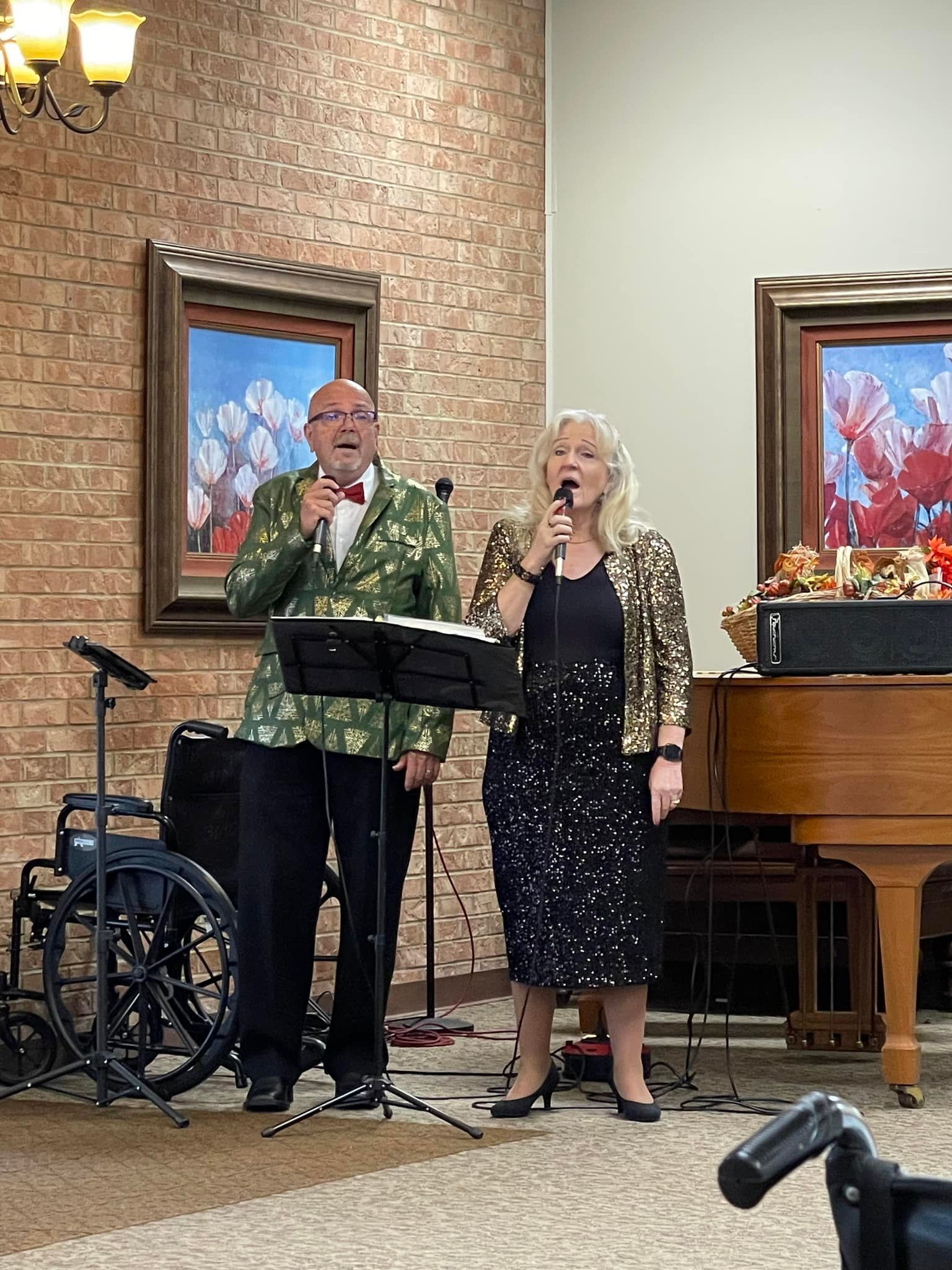 Two people singing with microphones on a stage. The woman in a sparkly outfit, the man in a green blazer.