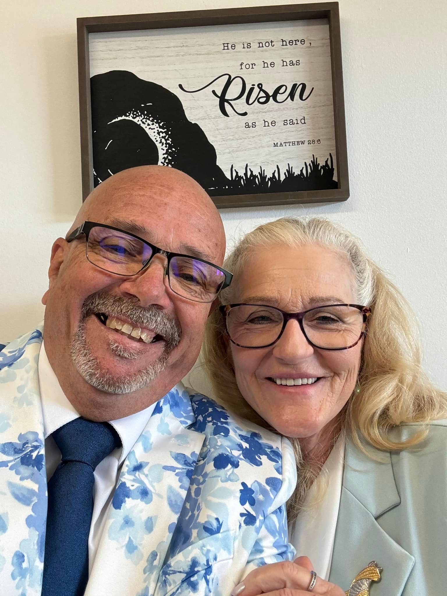 Smiling couple in front of a wall decor, man in blue floral suit, woman in light blue jacket.