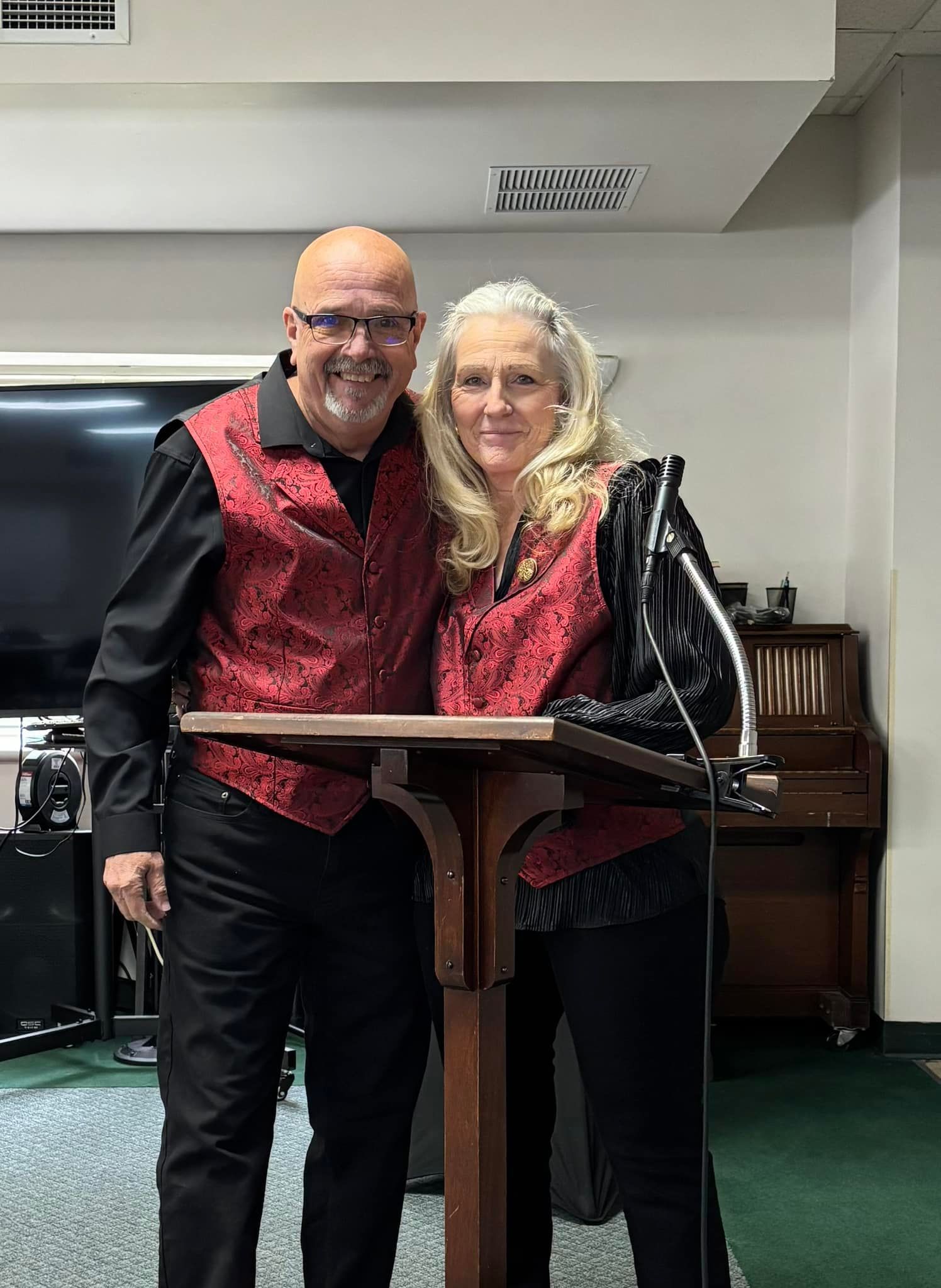 A man and woman in matching vests stand near a podium, smiling. Interior setting.