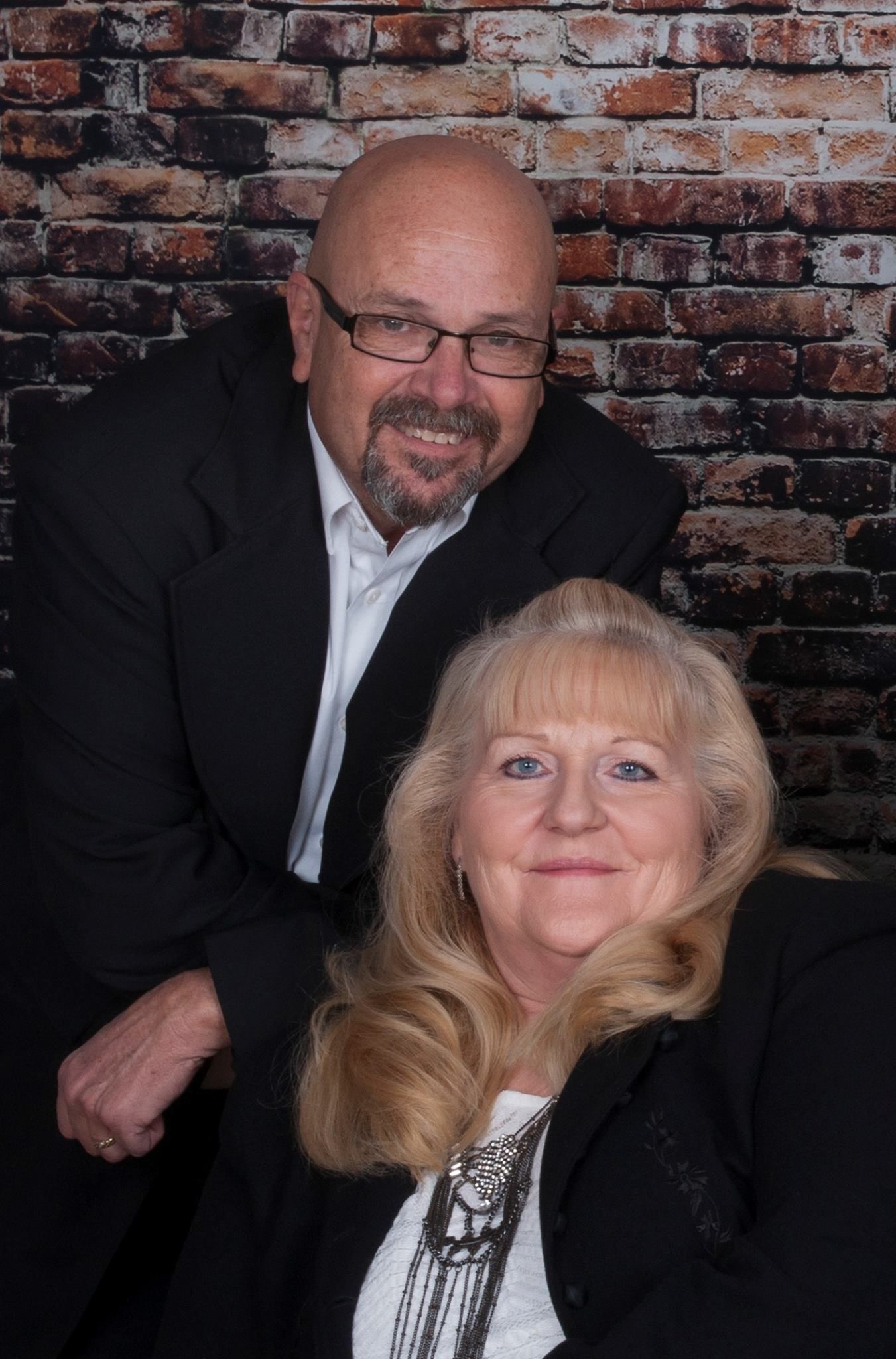 Couple, man with glasses in a suit behind woman with blonde hair, smiling in front of a brick wall.