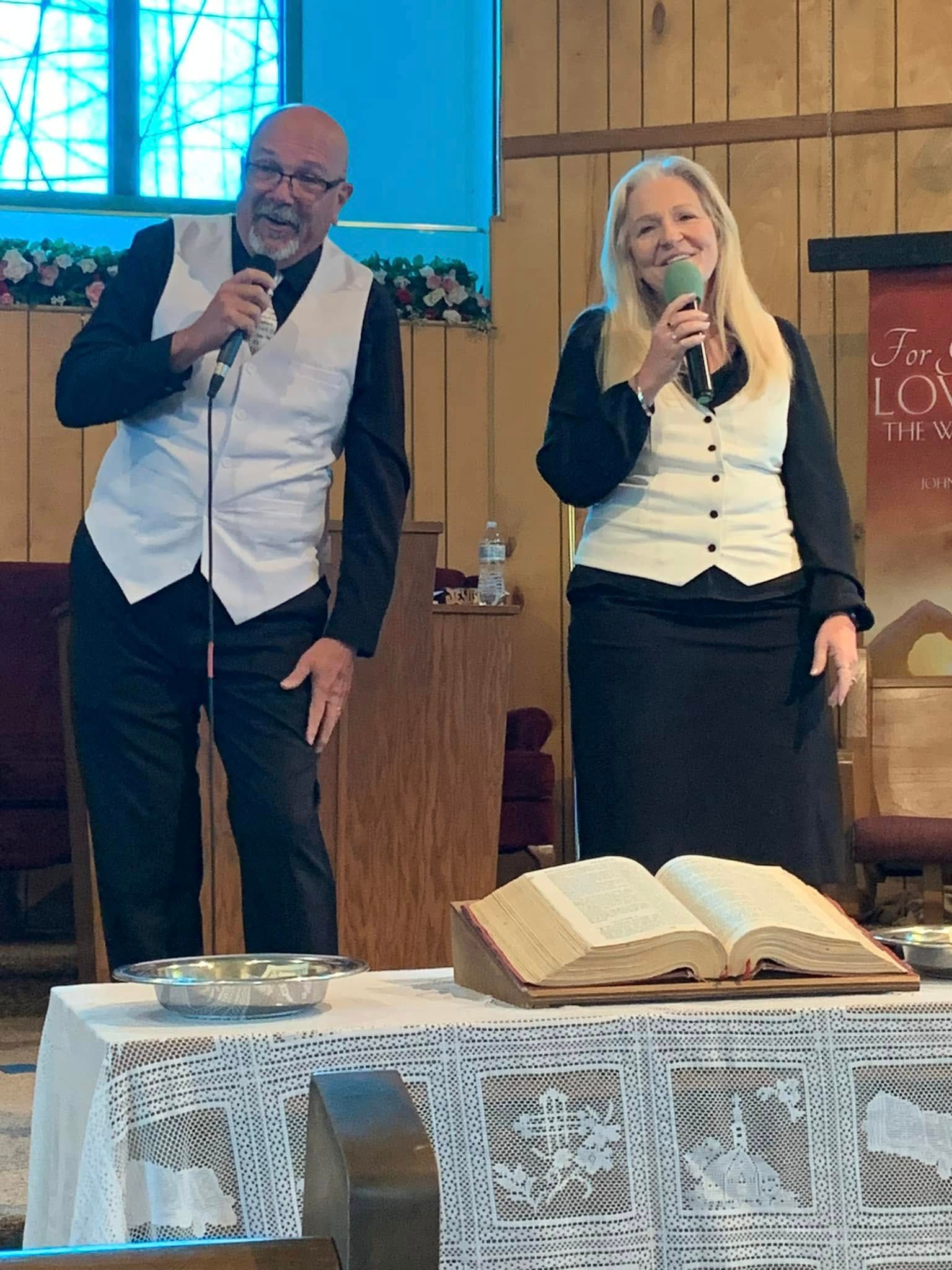 Two people singing in church, holding microphones, standing near an open Bible.