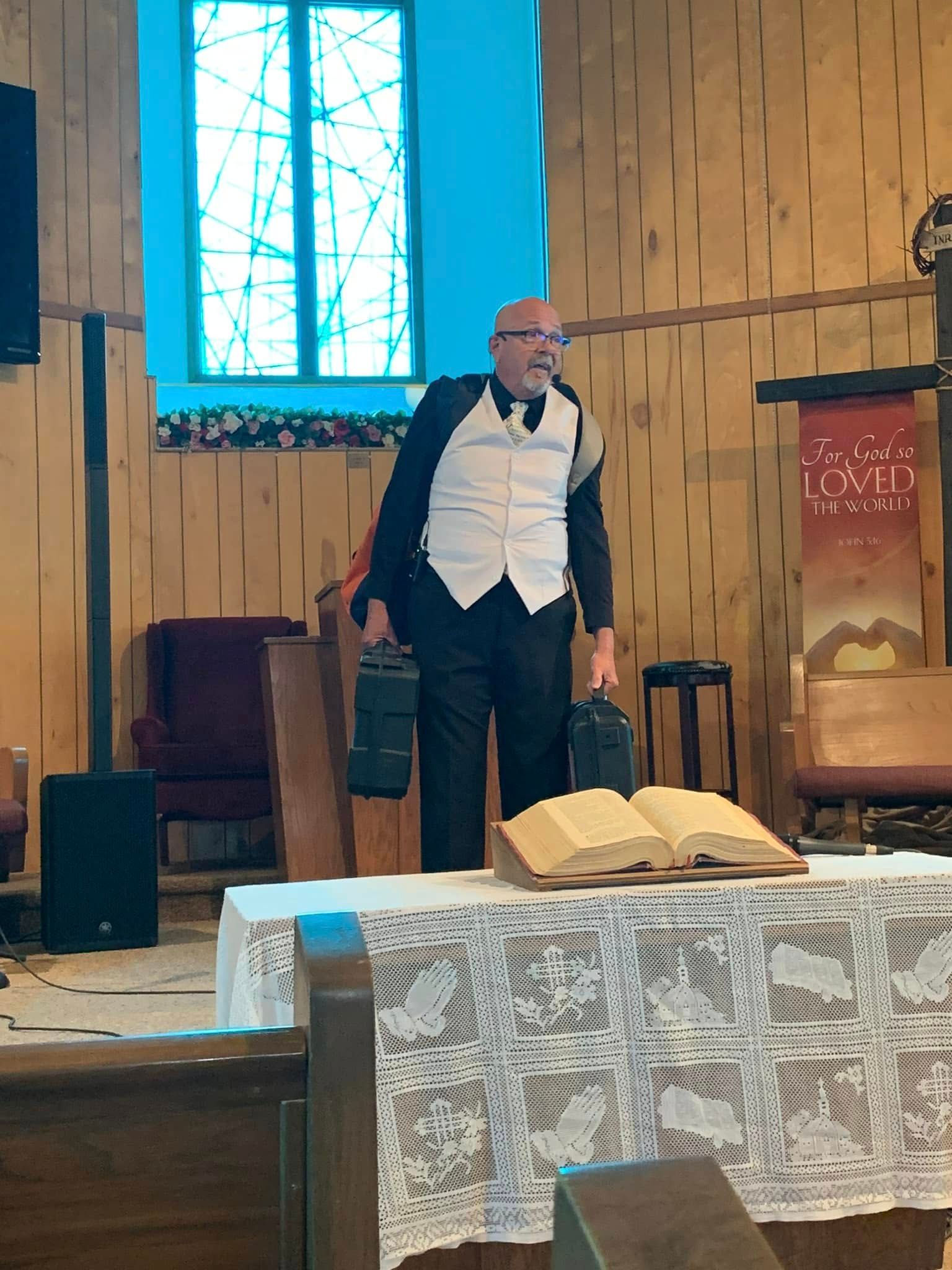 Man in vest holding two lights, speaking at pulpit with open book in church.