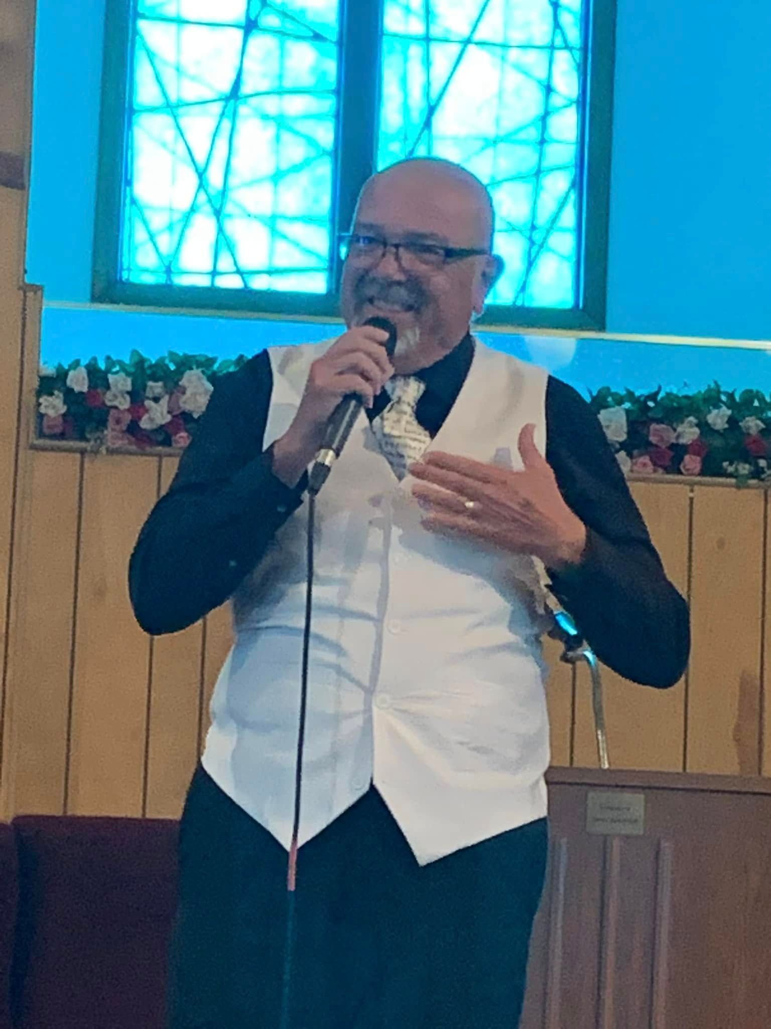Man with a shaved head, vest, and microphone speaking in front of a stained-glass window.
