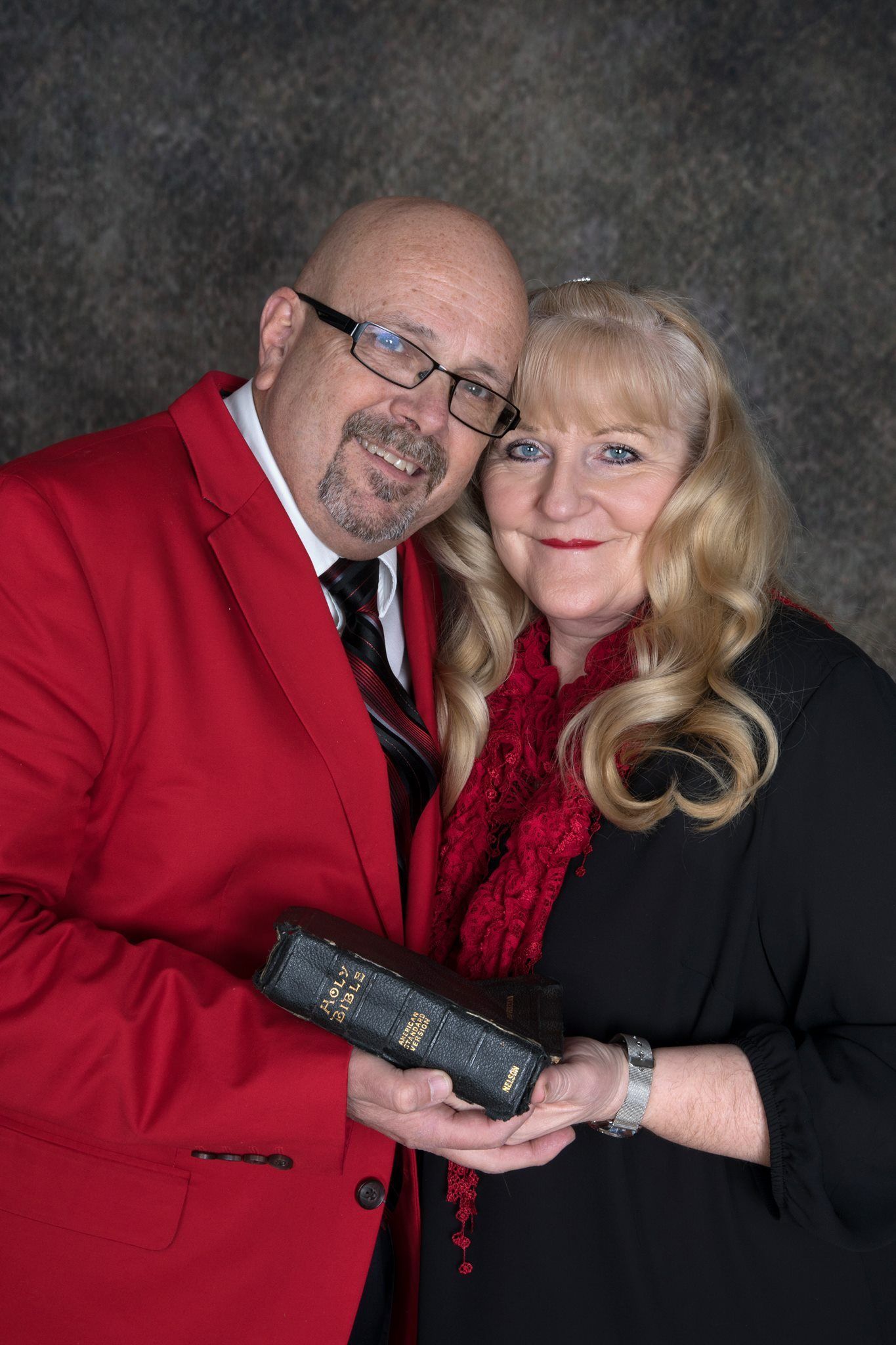 Couple holding a Bible; man in red blazer, woman in black; studio portrait with a gray background.