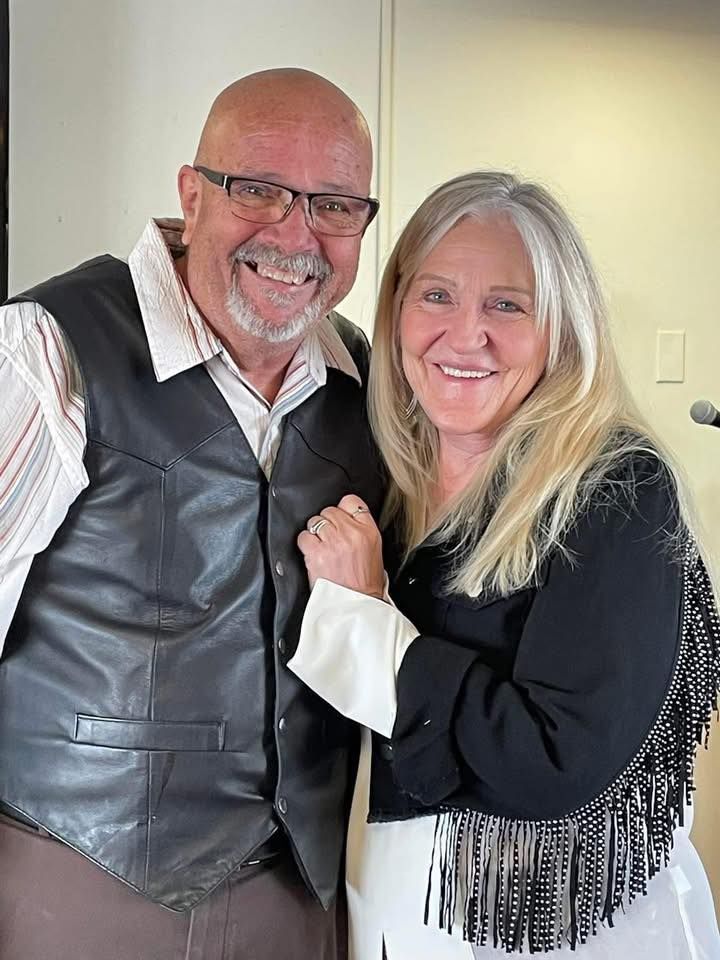 Man and woman smiling, posed closely together. Man wears glasses, vest; woman has long blonde hair, fringe.