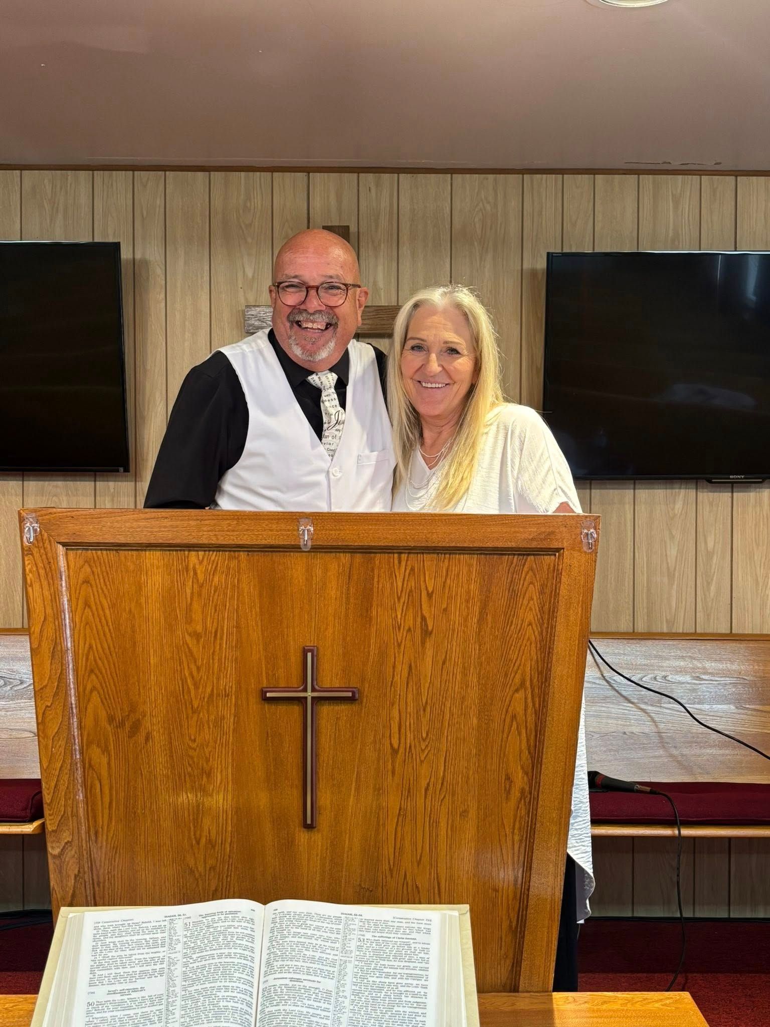Man and woman standing behind a church pulpit, smiling. Man wears glasses, vest, and tie. Woman in white dress.