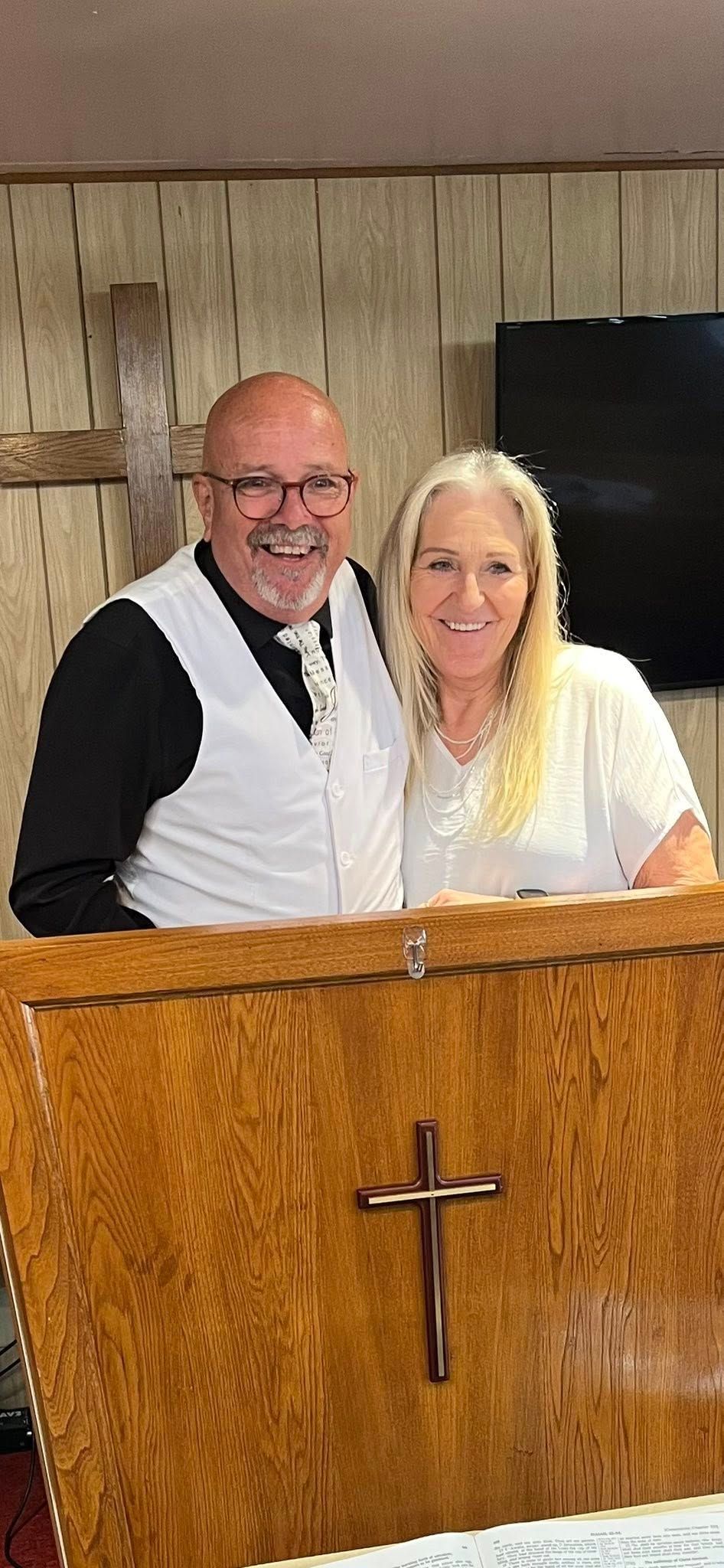 A smiling man and woman stand behind a wooden podium with a cross.