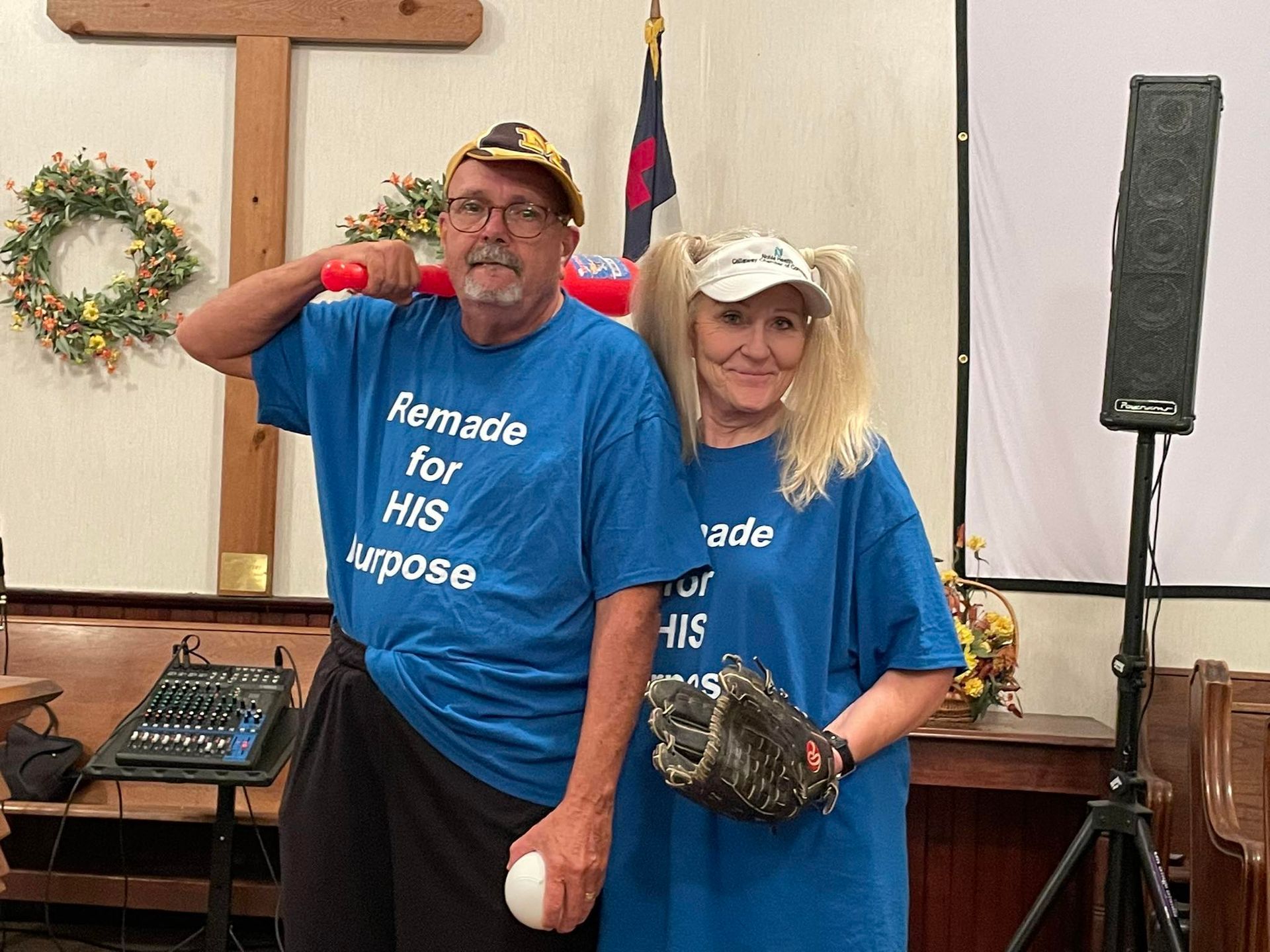 A man and woman in blue shirts, holding baseball gear, in a church, with a cross in the background.