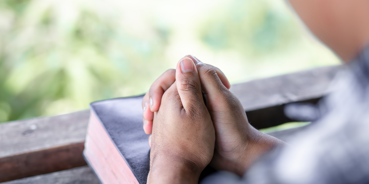 A person is praying with their hands folded over a bible.