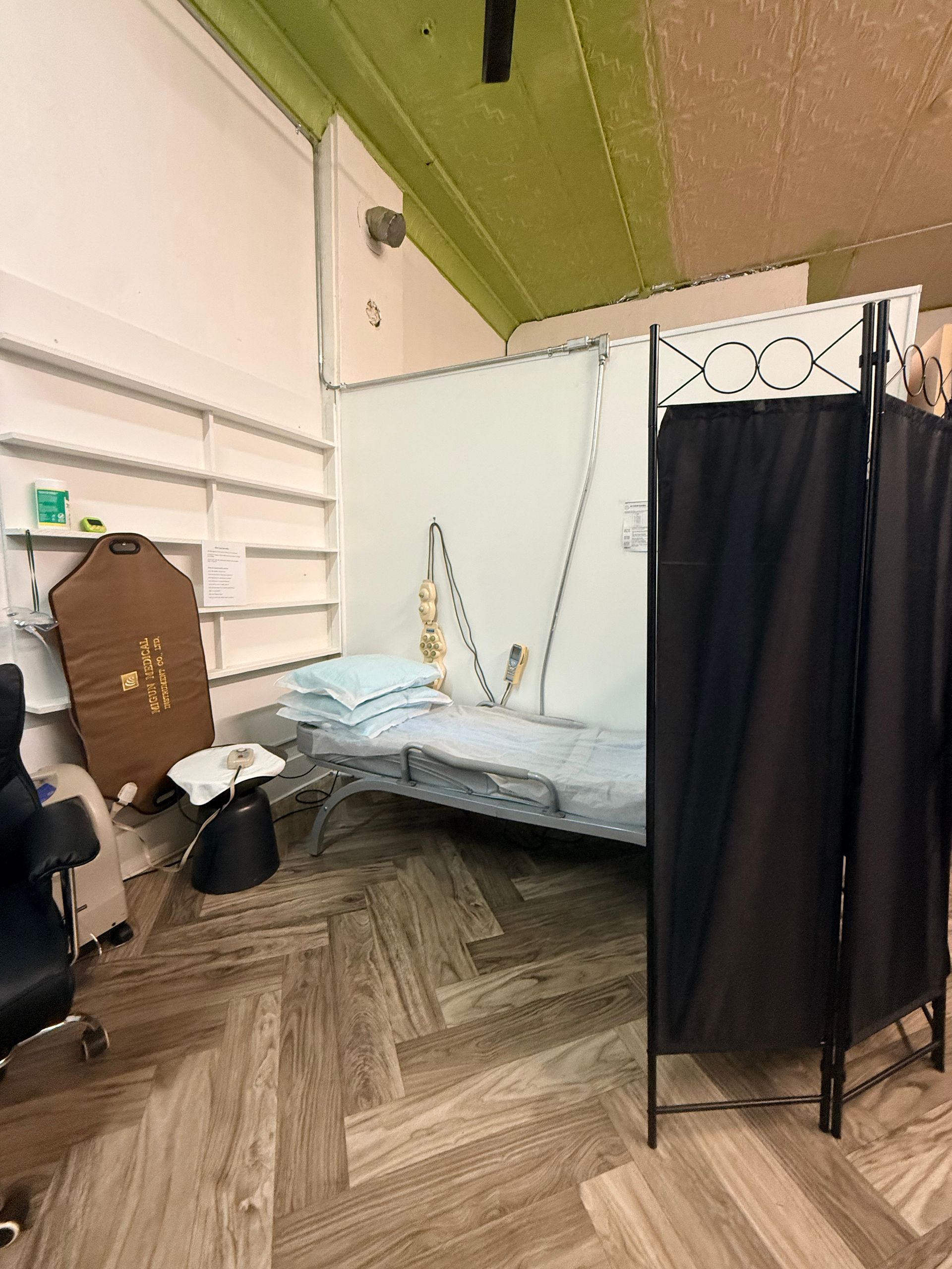 A medical examination area with a treatment bed, a privacy screen, and a stool on a herringbone wood-patterned floor.