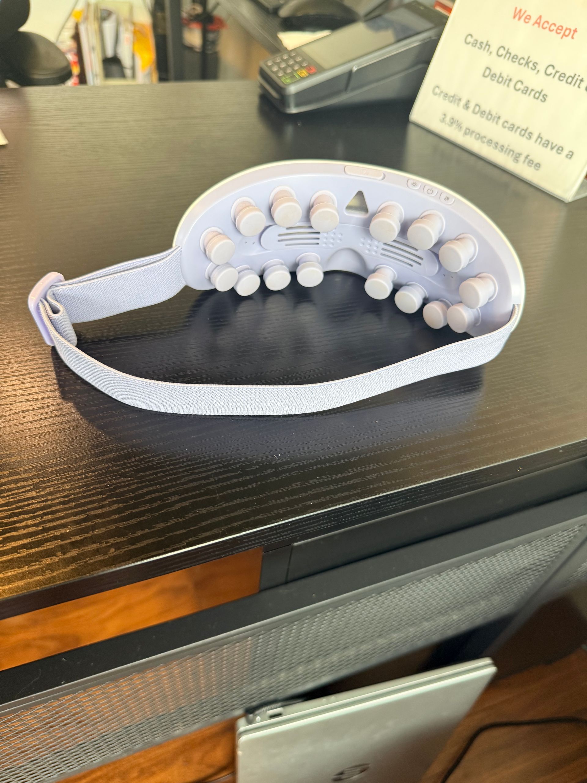 A lavender-colored eye mask with massage nodes on the interior sits on a dark desk next to a credit card terminal.