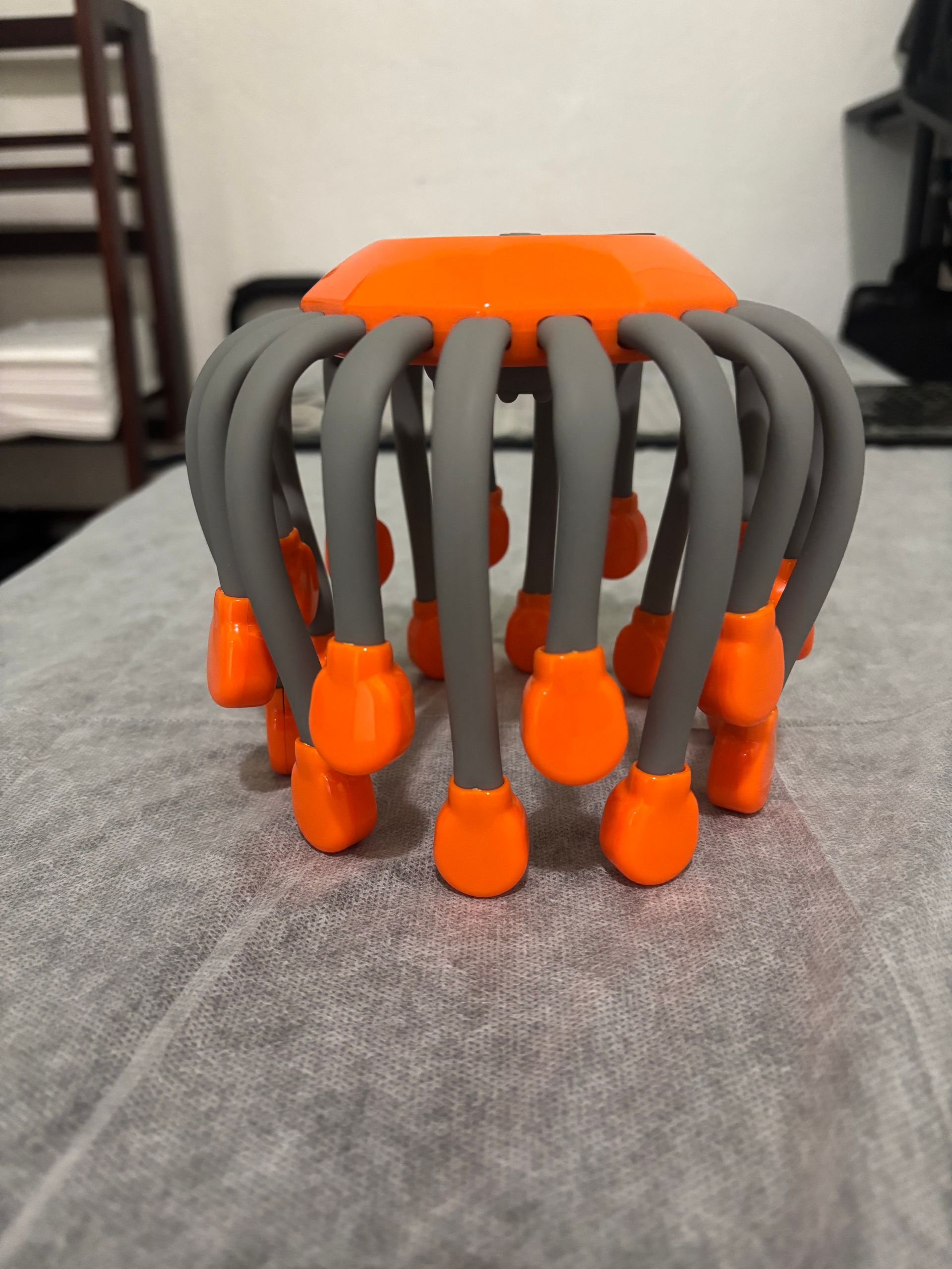 A handheld scalp massager with a bright orange handle and multiple gray wire prongs ending in small orange tips.