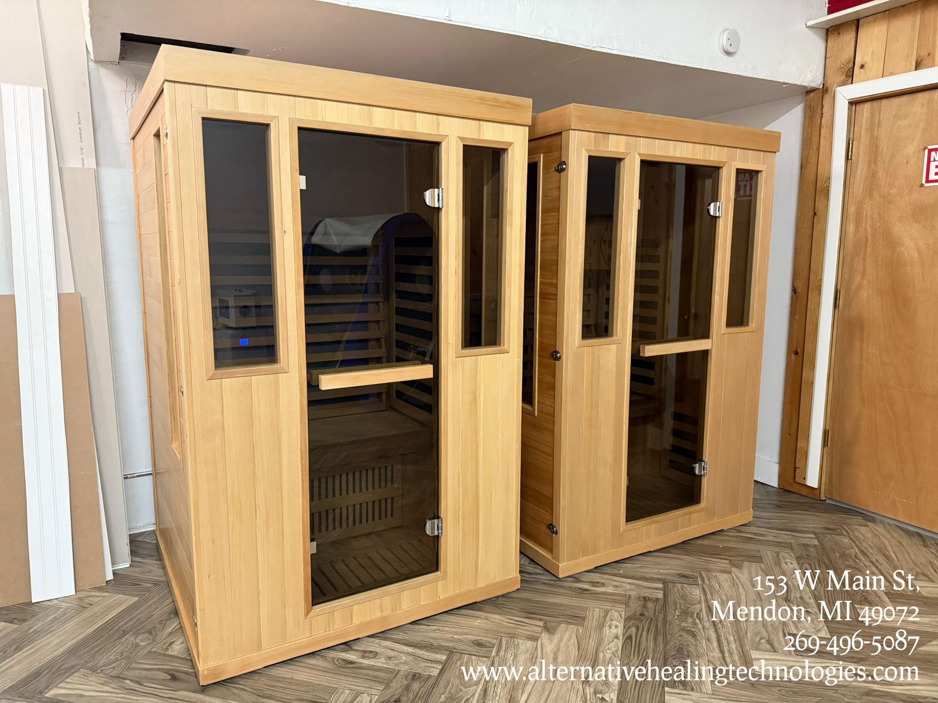 Two wooden saunas with glass doors and windows sit in a room, ready for use.