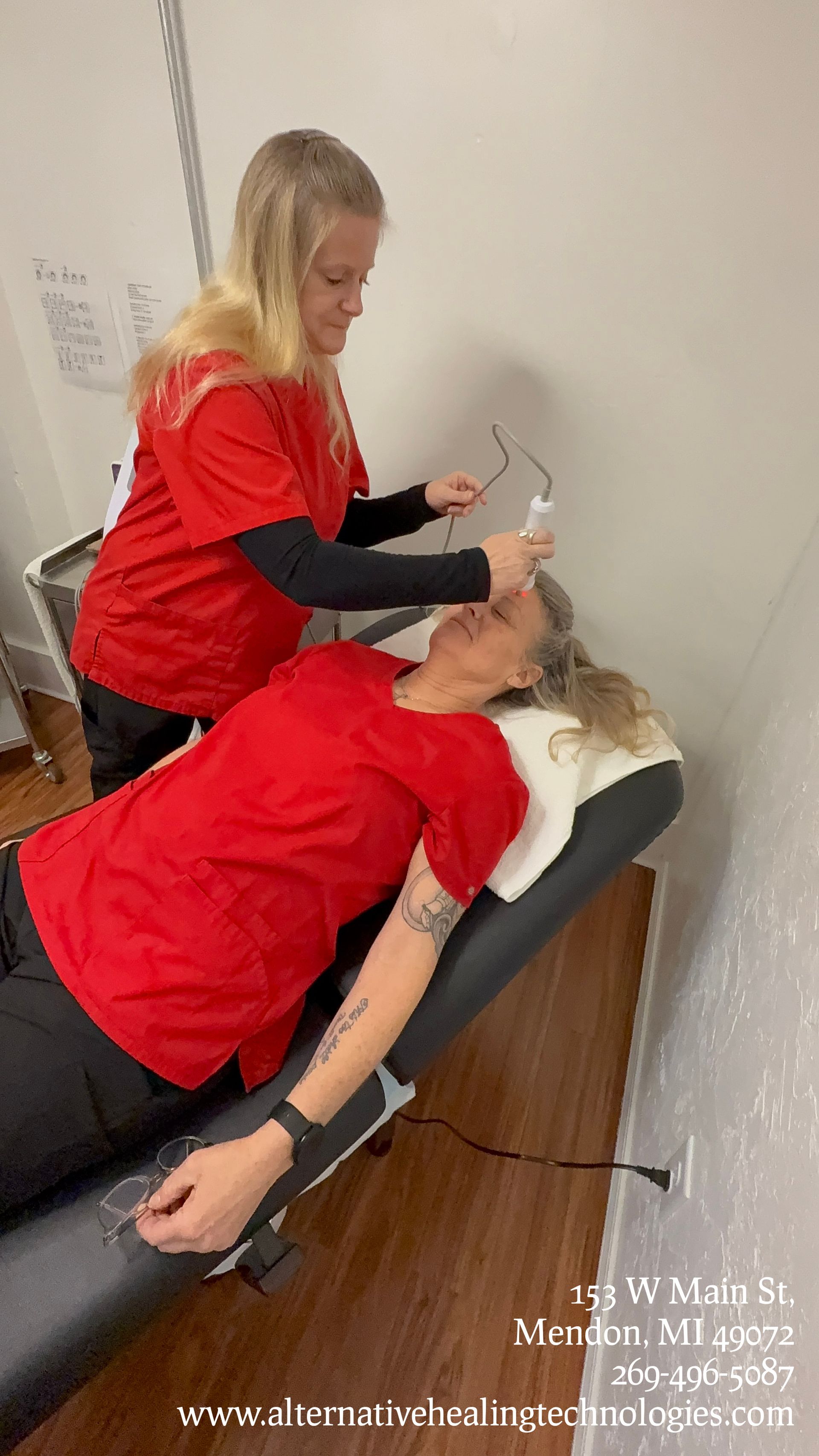 A practitioner in a red uniform uses a handheld device on the forehead of a client lying on a treatment table.