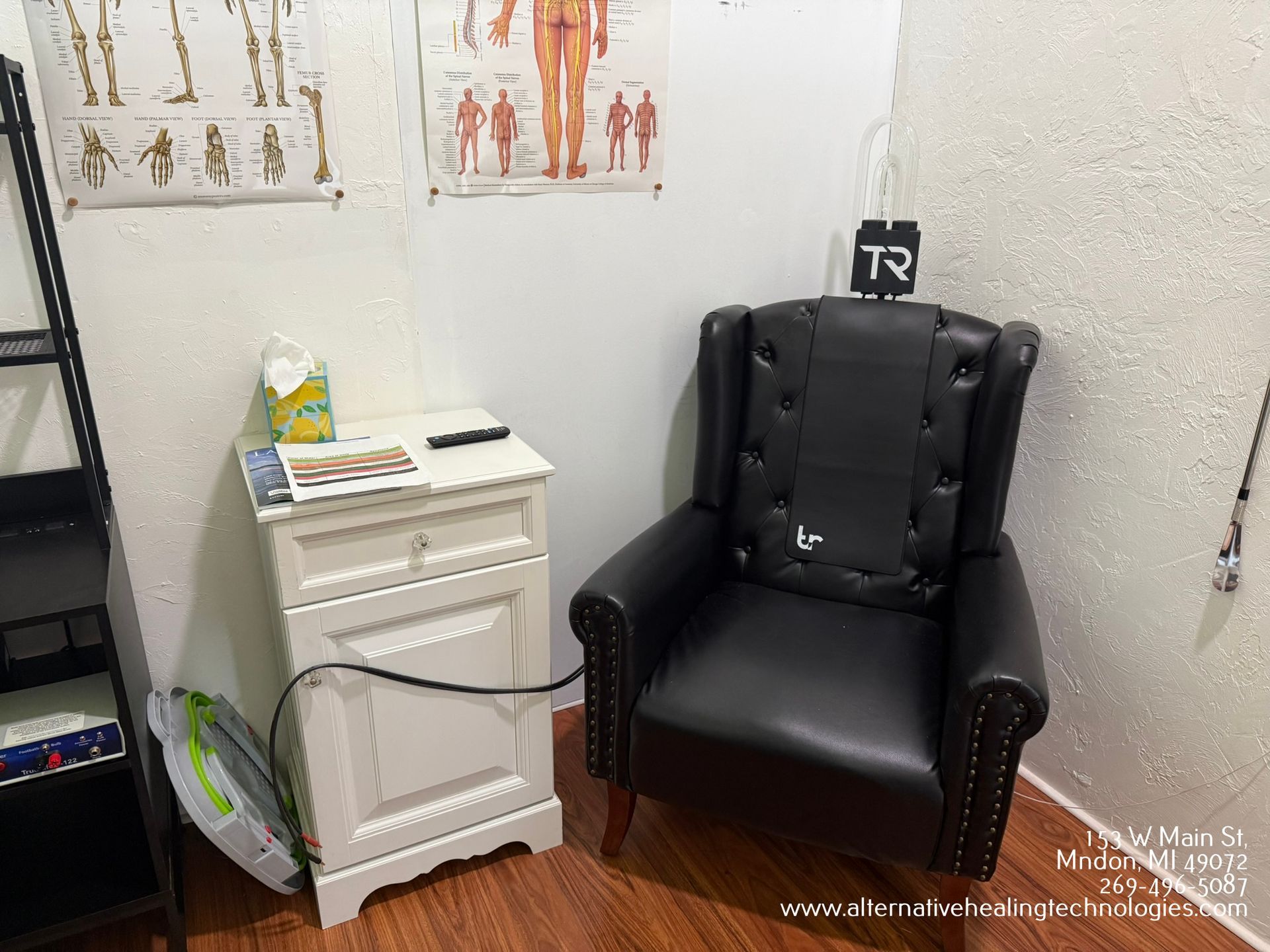 A black armchair in a corner next to a white nightstand in an office. Anatomical charts hang on the wall.