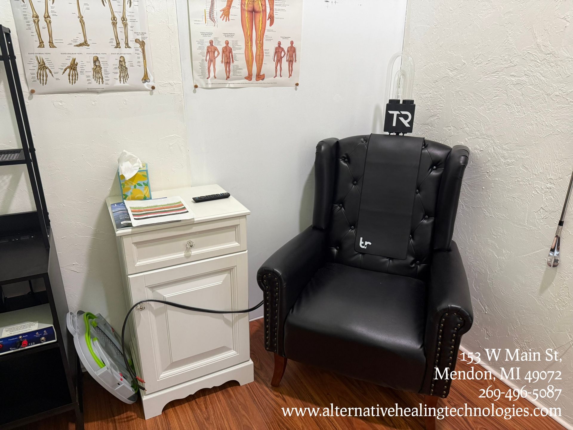 A black armchair in a corner next to a white nightstand in an office. Anatomical charts hang on the wall.