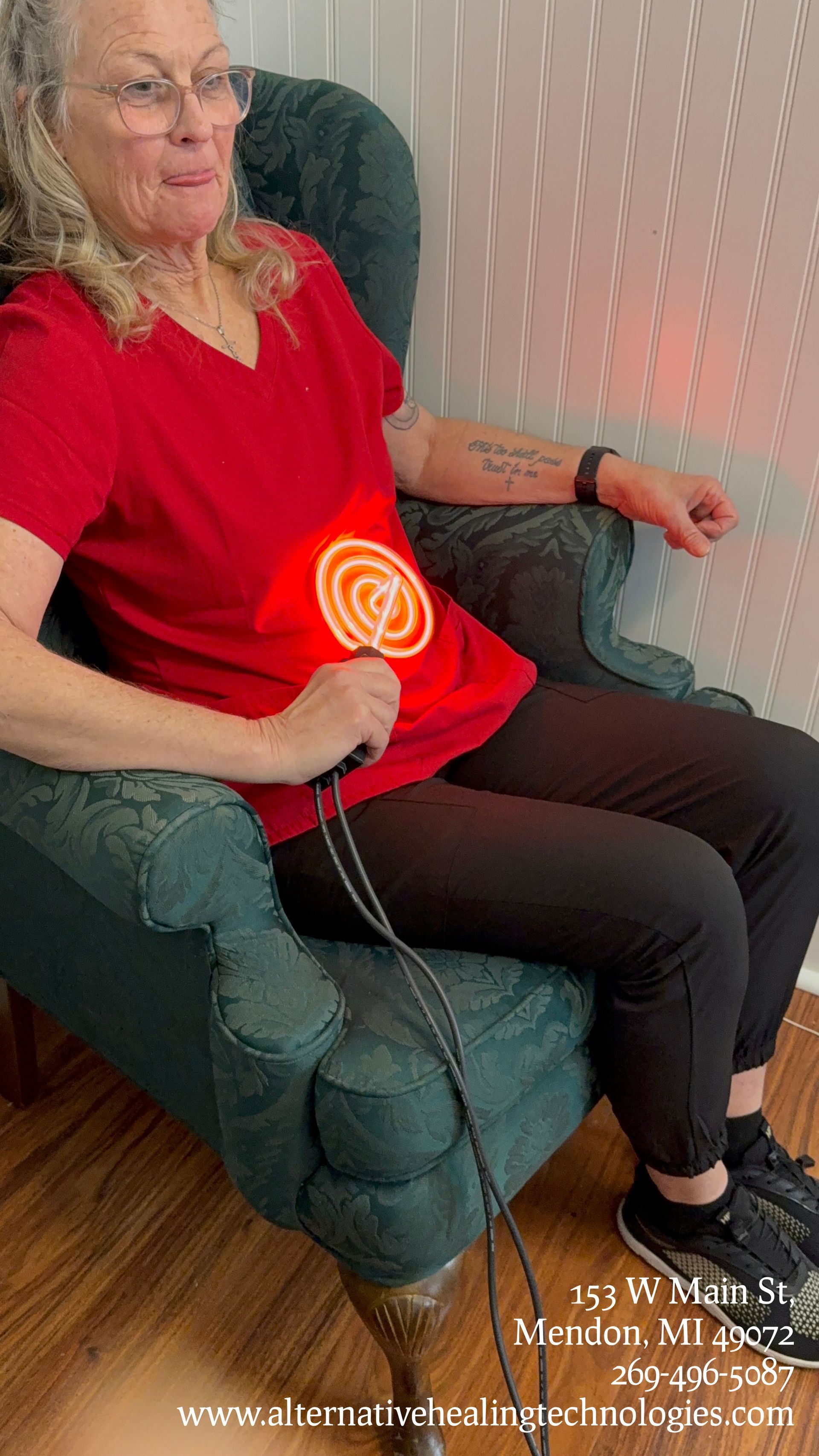 Woman in red shirt using a red light therapy device on her stomach while seated in a green armchair.
