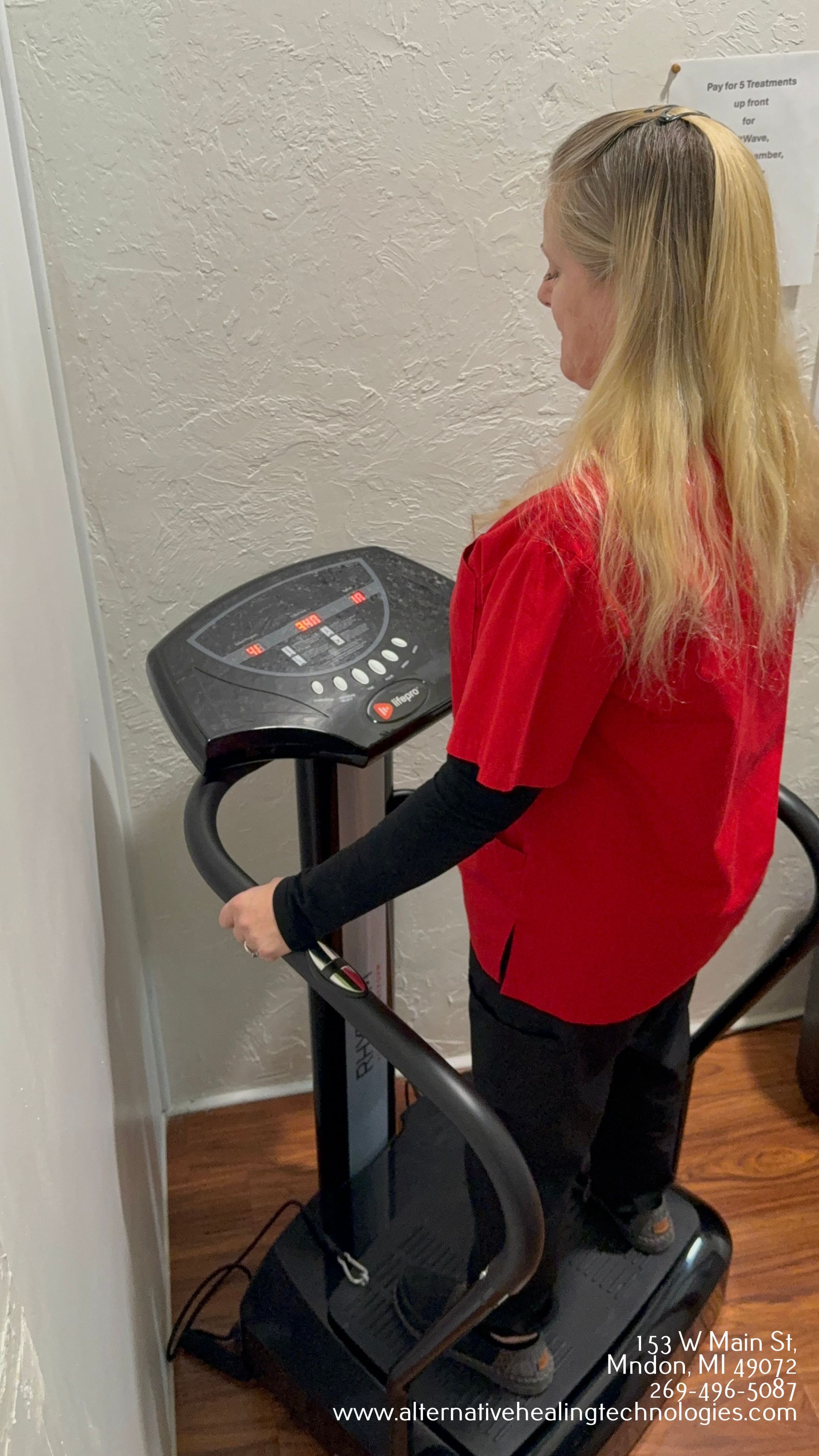 Woman using a vibrating exercise machine, indoors, wearing a red shirt, smiling.