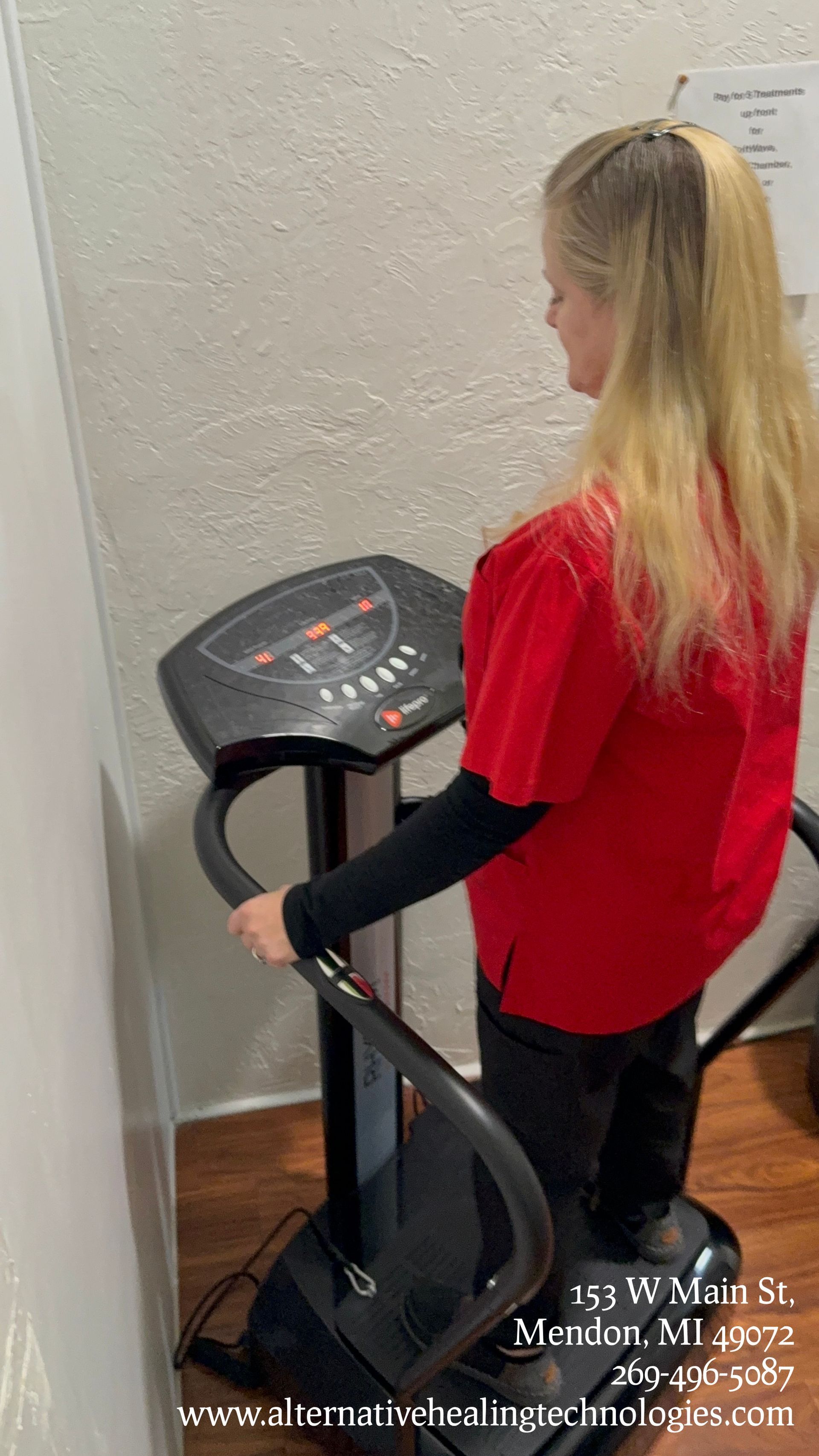 Woman in red shirt on a vibrating exercise machine, smiling. Indoor, near a wall.
