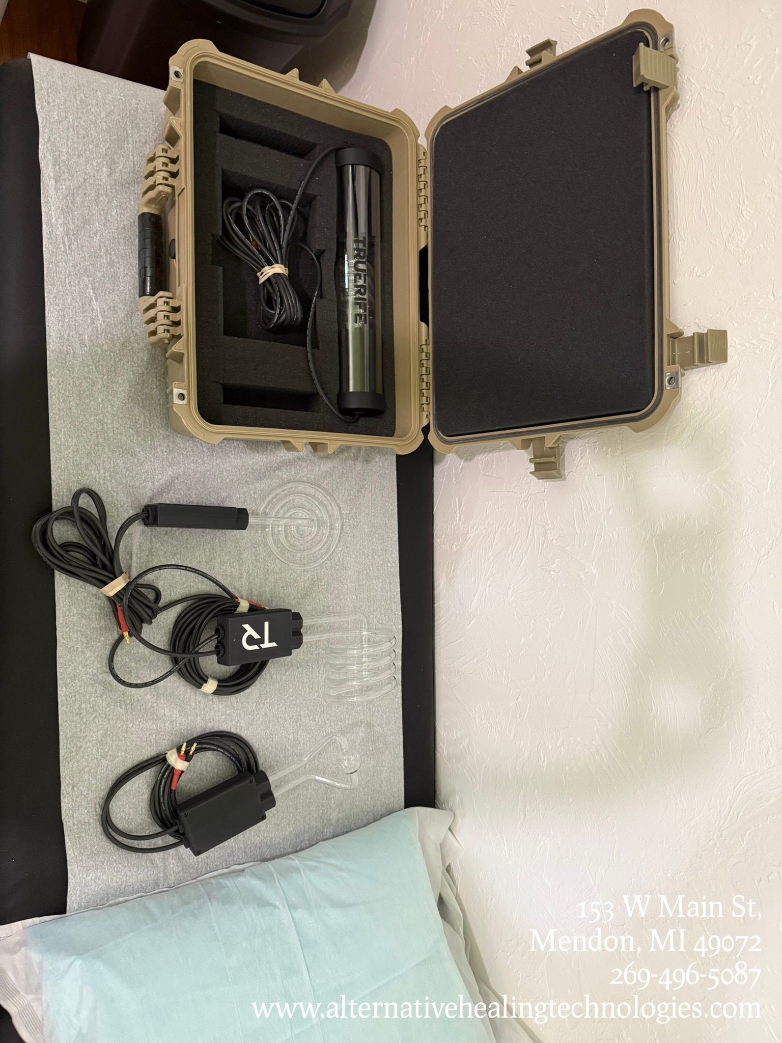 A treatment room with a massage table, equipment, and a case. A trash can and chair are nearby.