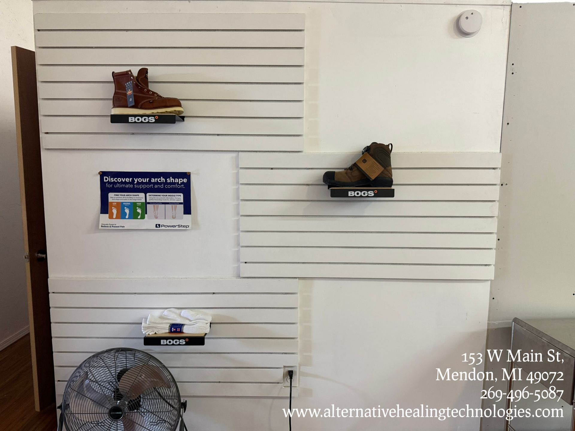 A display wall with white shelving, three Ecco shoes on brackets, an informational sign, and a fan, located in Mendon, MI.