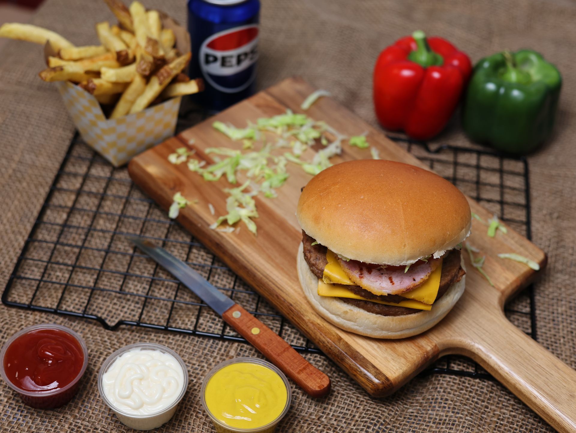 Hamburger with cheese and ham on a wooden board, fries, Pepsi, peppers, and condiments.