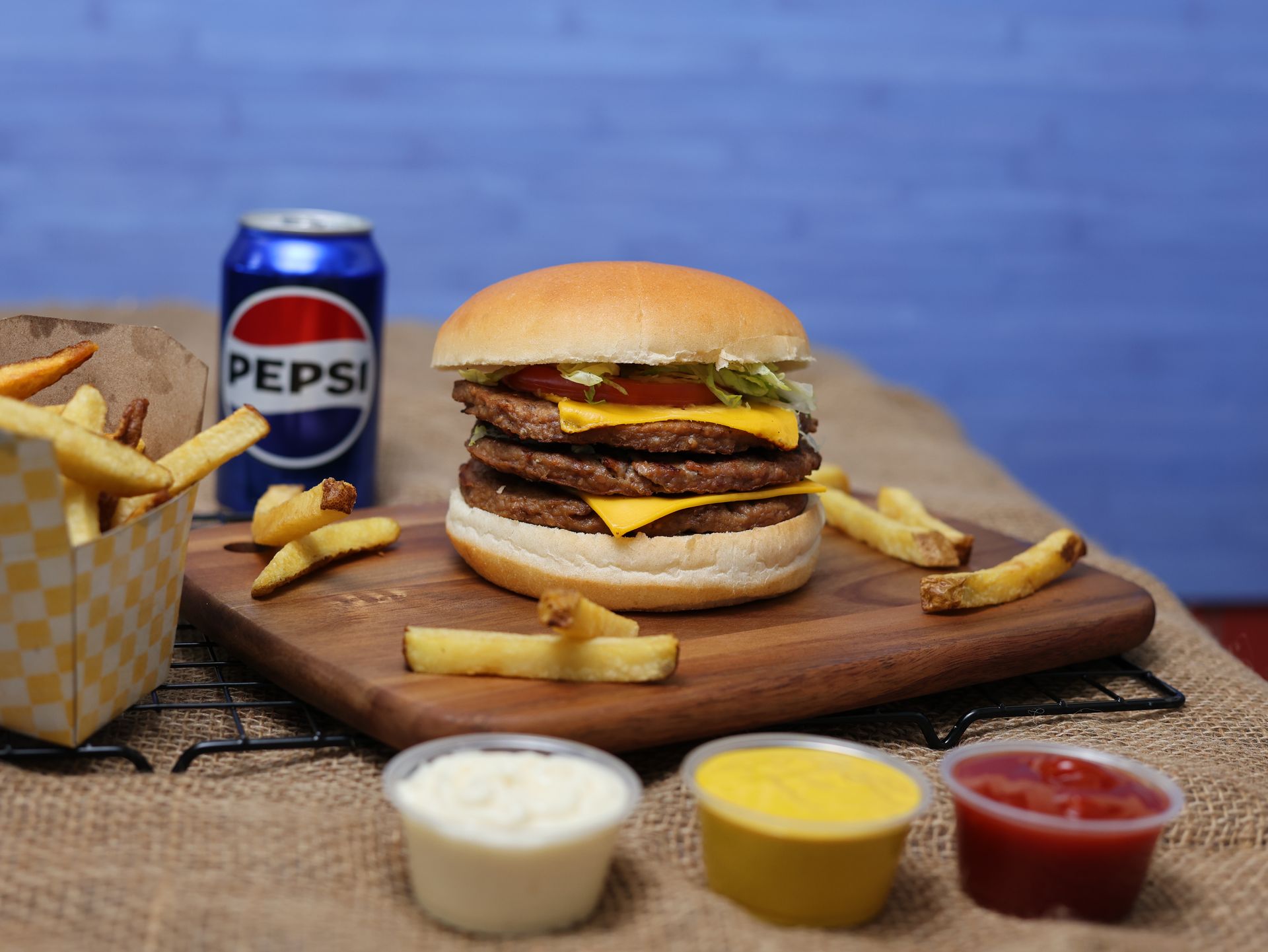 Double-decker burger with cheese, fries, Pepsi can, and dipping sauces on a wooden board.