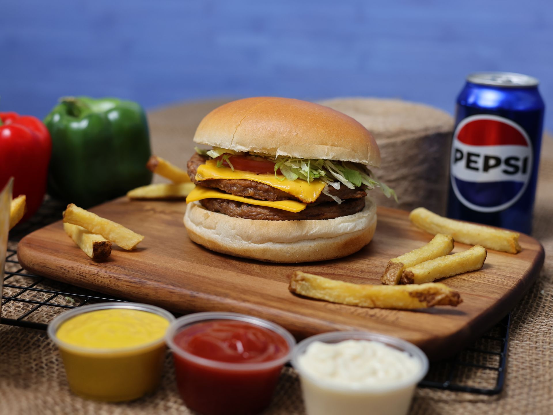 Double cheeseburger, fries, and Pepsi on a wooden board with condiments and bell peppers.