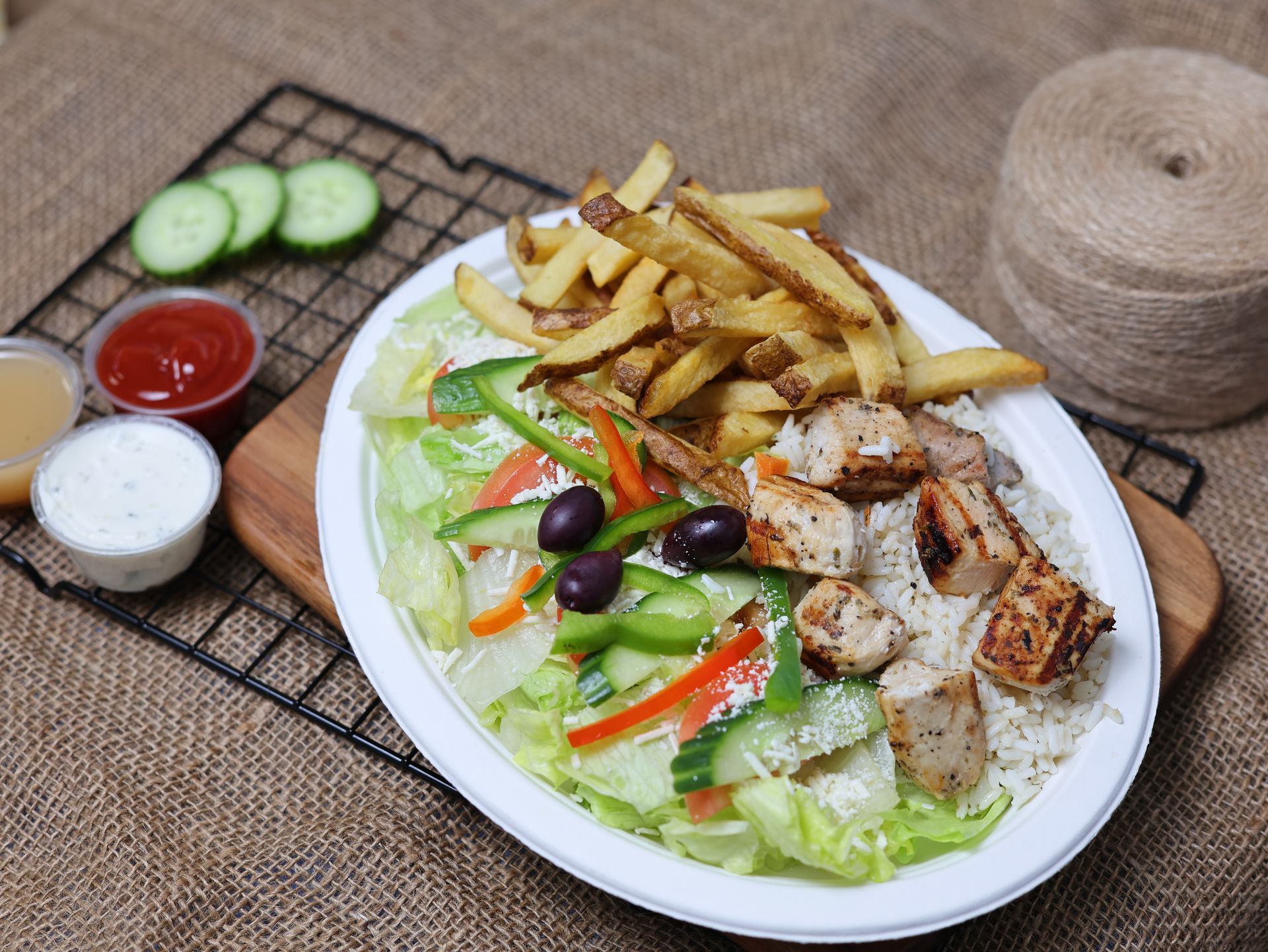 Plate of grilled chicken, rice, fries, and salad, with dipping sauces and sliced cucumbers.
