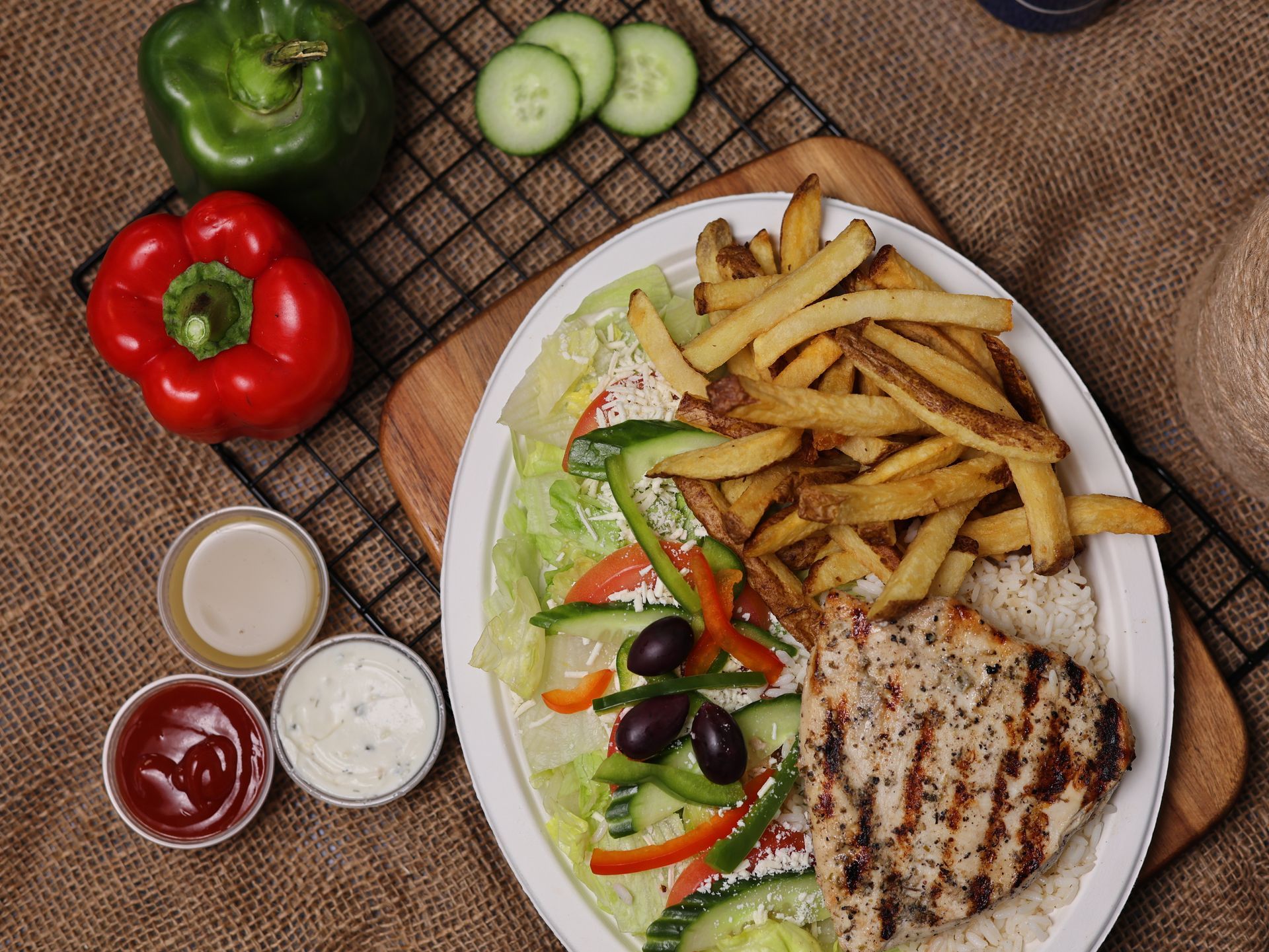Grilled chicken and fries plate with salad and sauces, ingredients arranged on rustic surface.