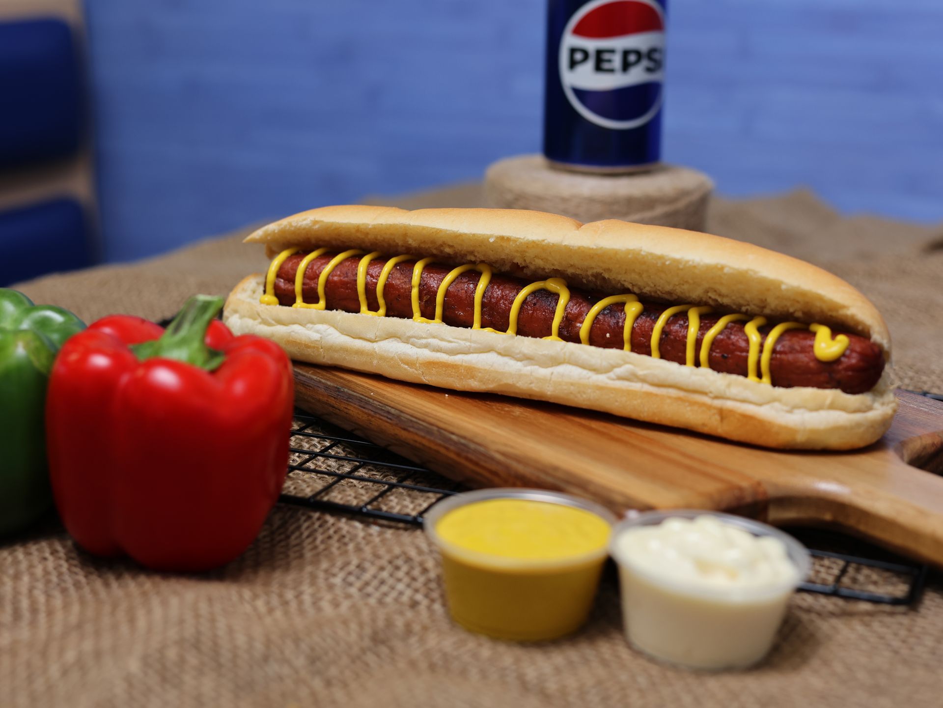 Hot dog with mustard, peppers, and Pepsi can on a wooden board.