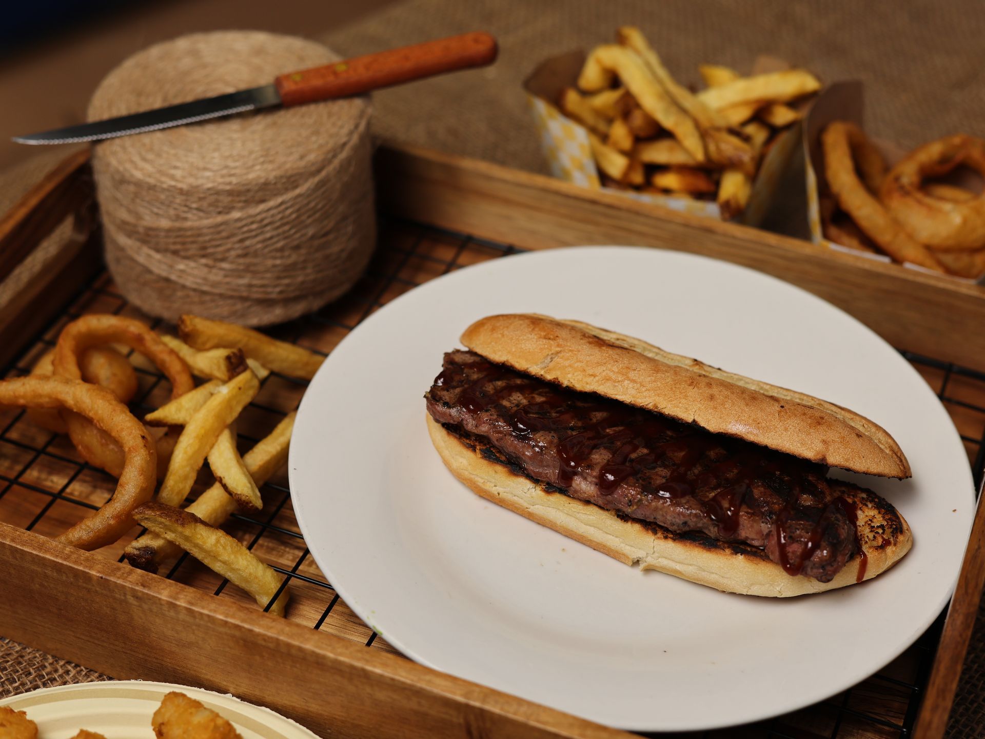 Steak sandwich on a white plate with onion rings and fries in a wooden tray.