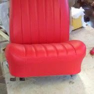 Red upholstered chair seat with vertical stitching and black legs in a workshop setting