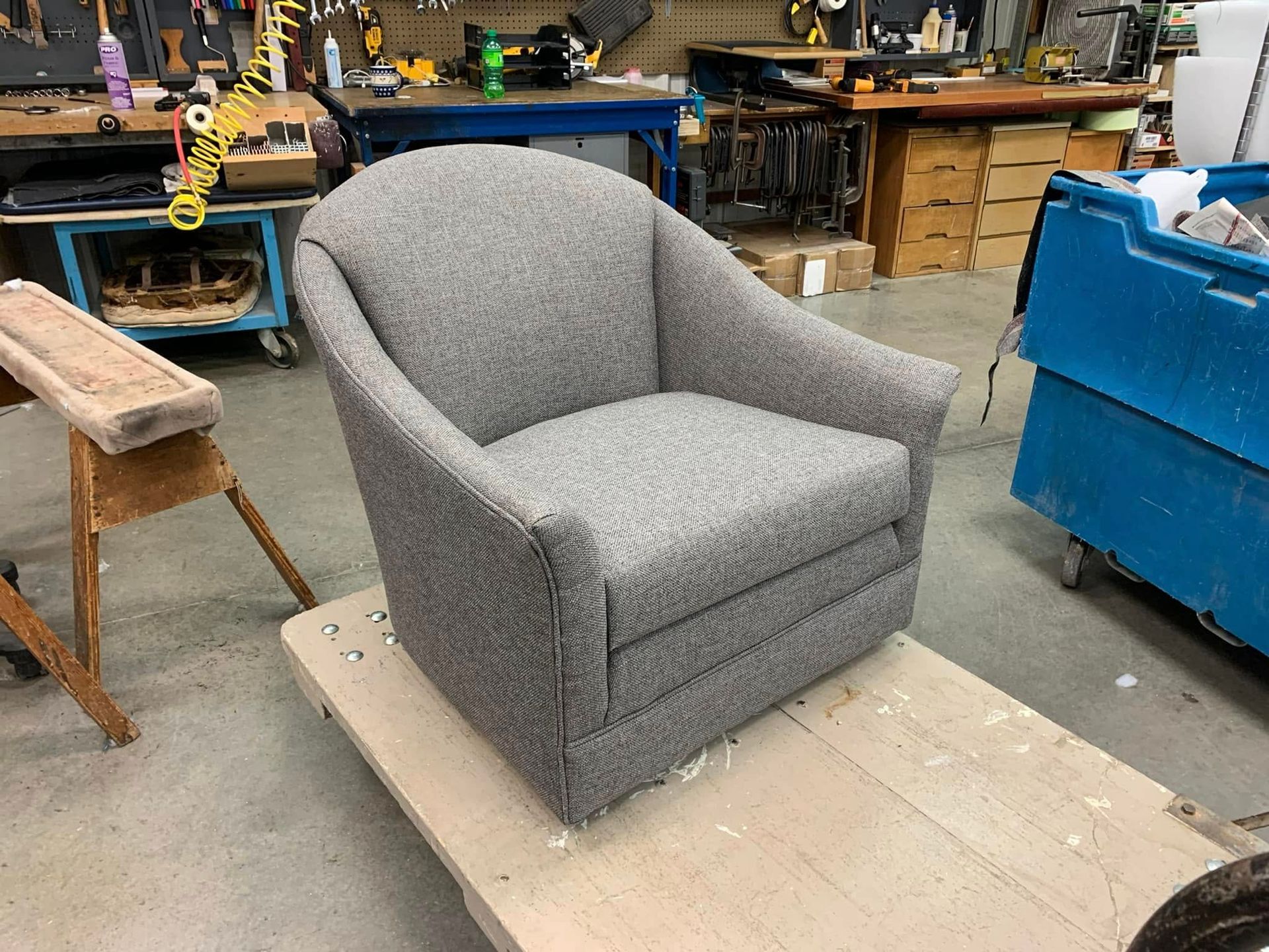 Gray upholstered armchair on a plywood platform in a workshop, surrounded by tools and benches