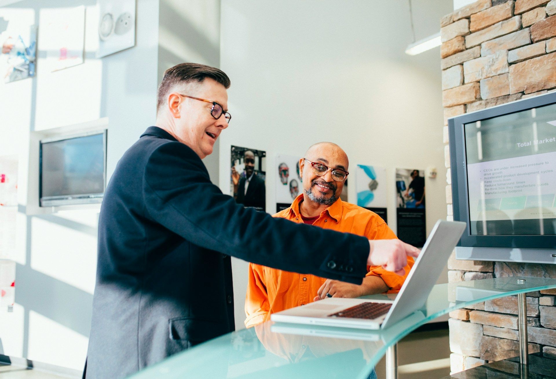 A man is pointing at a laptop computer while another man looks on.