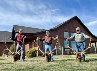 Three men are standing in front of a house holding elk antlers.