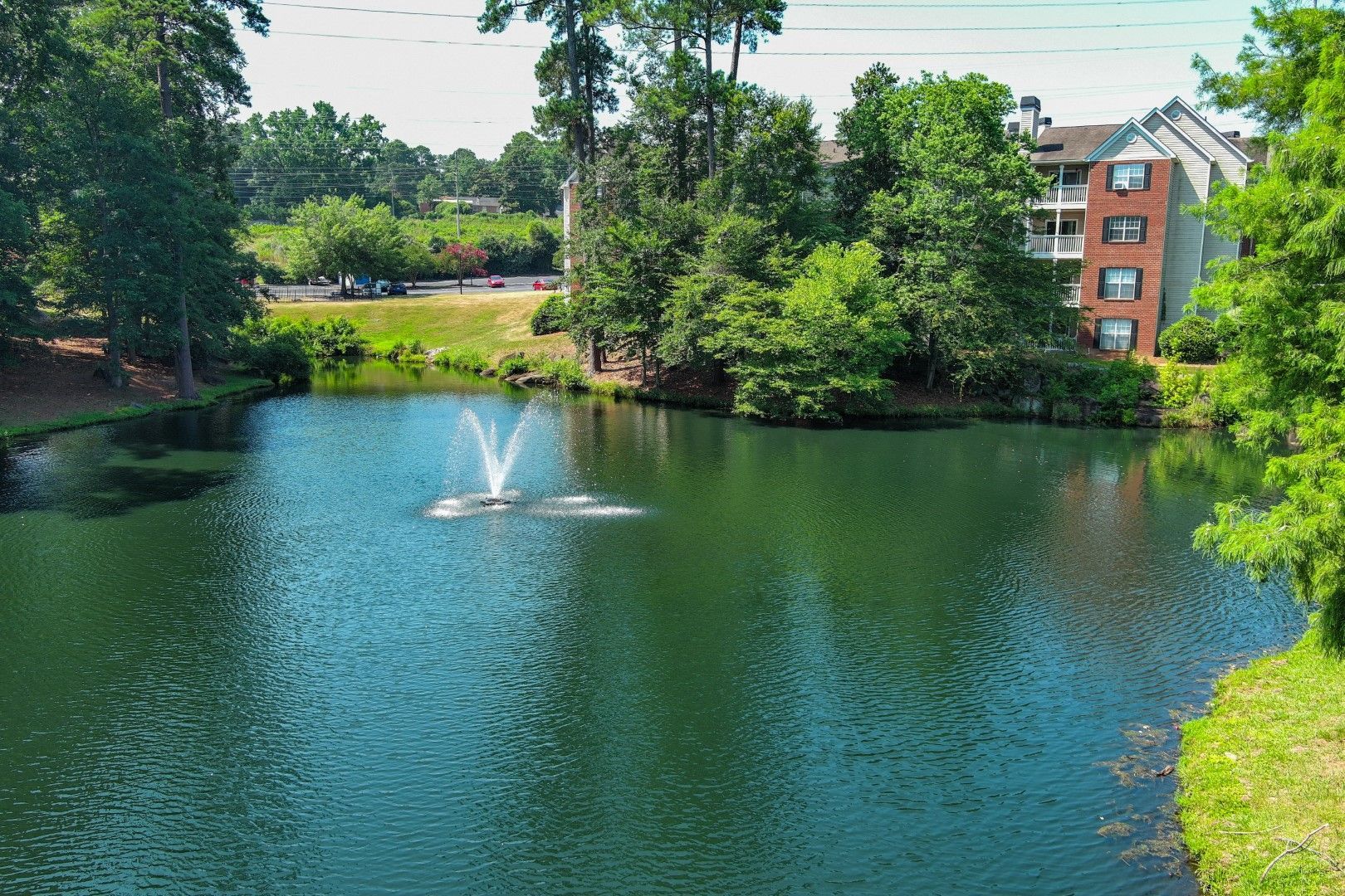 Pond with fountain, trees, and apartment building in background. Green water and lush greenery on a sunny day.