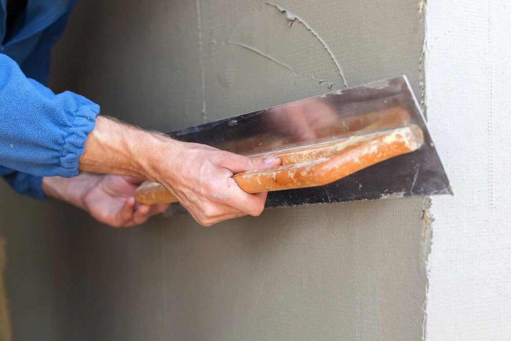 A Close-up View Of A Hand Using A Metal Trowel To Smooth Gray Plaster Onto A Wall — HJK Plastering In Ballina, NSW