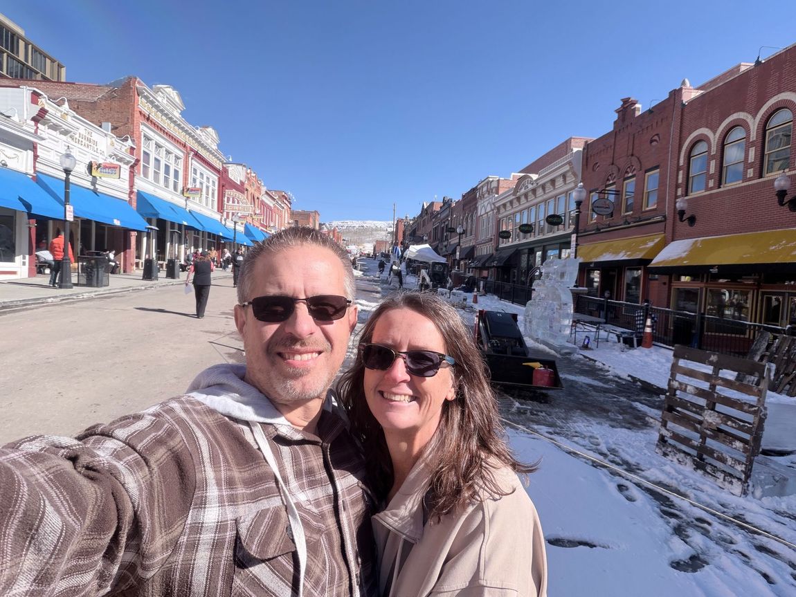 Smiling couple in sunglasses posing on a snowy Main Street lined with shops under a bright blue sky