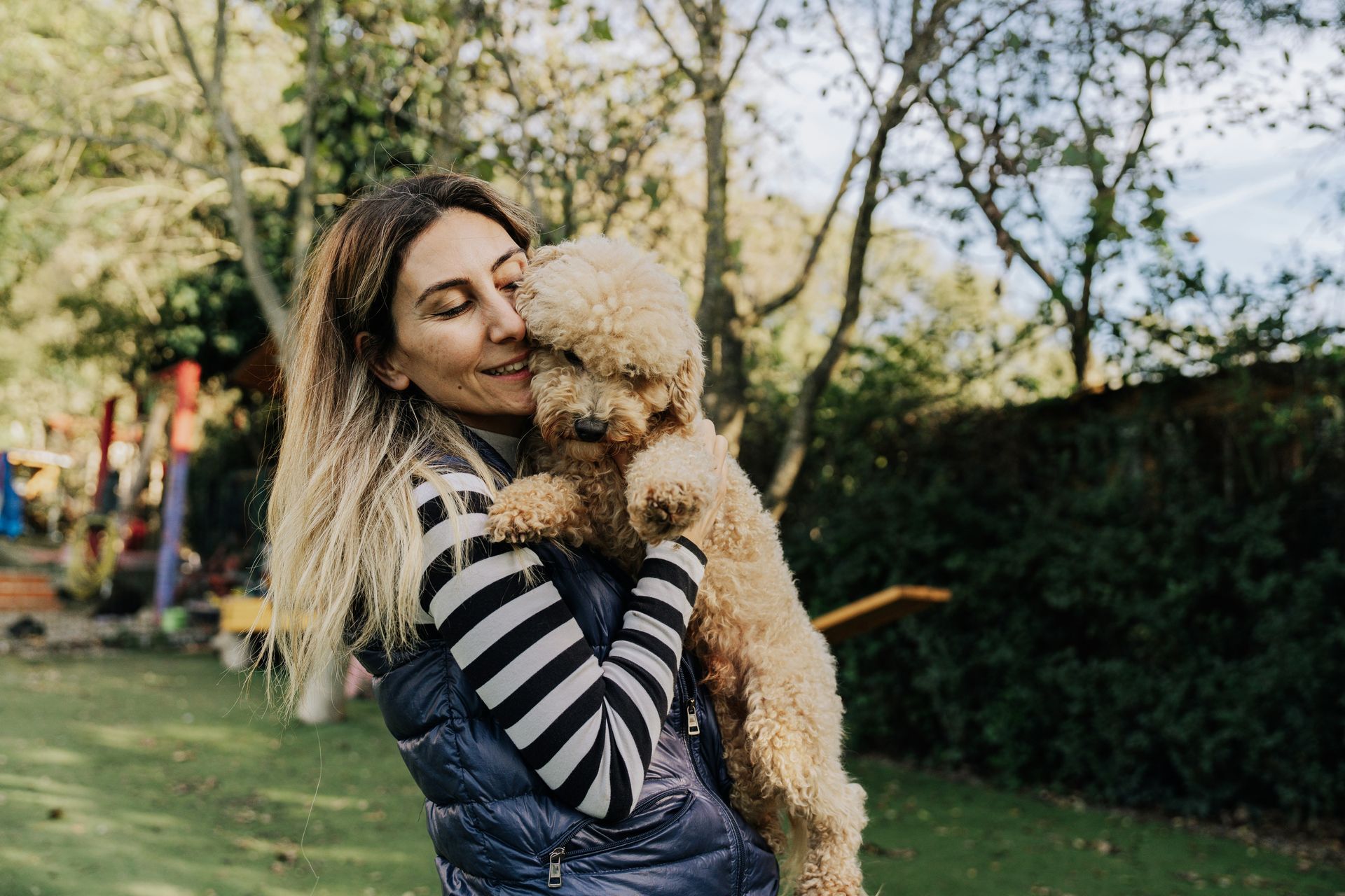 Woman hugging a curly-haired dog outdoors in a park. The woman is smiling, and the dog is cuddled close.