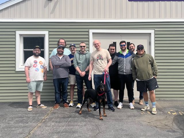 Group of eight people standing outside a beige building, with a black dog in front and one person holding a long object.