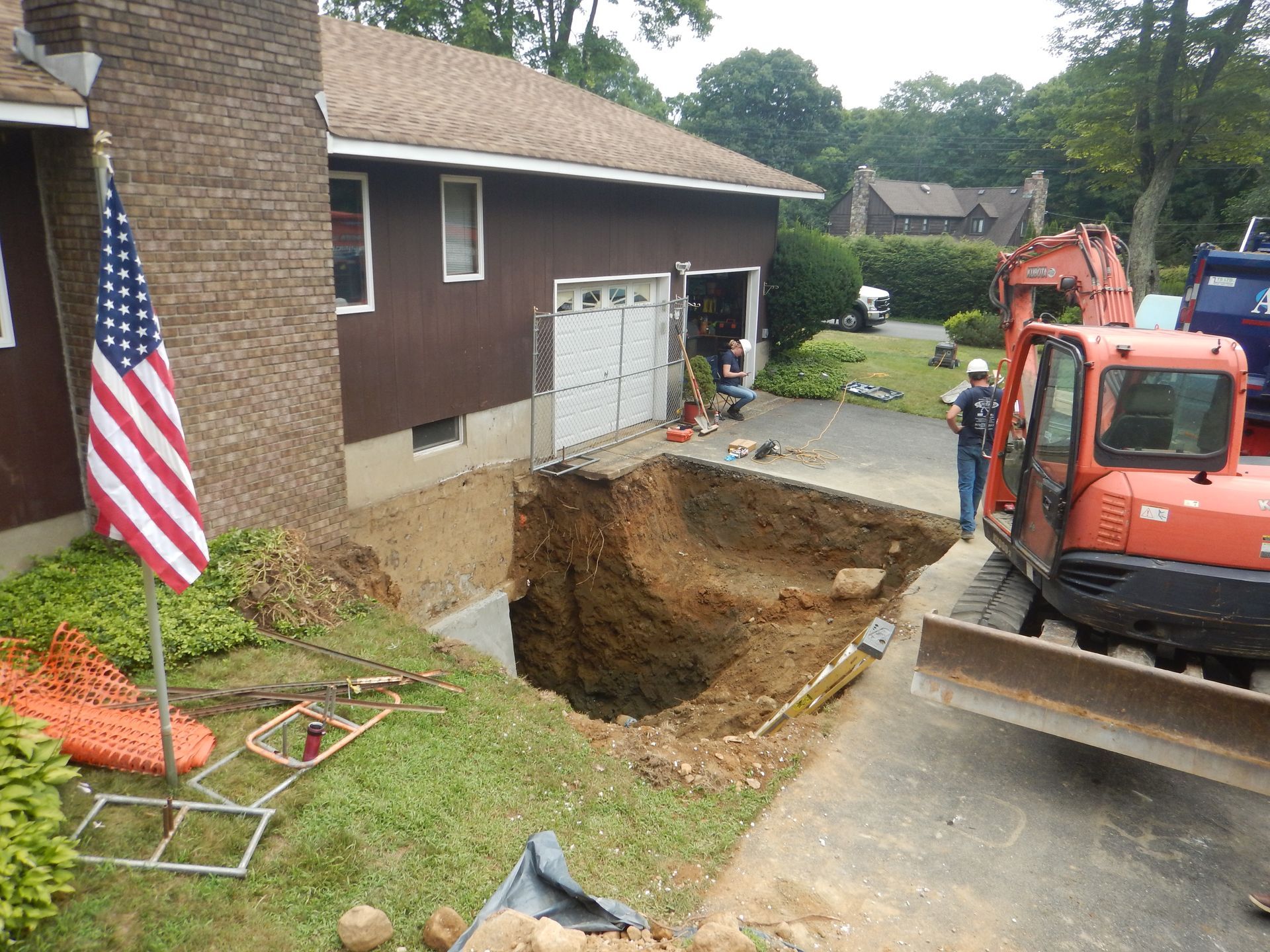 Construction site: Excavator digging a large hole near a brown house and garage with a US flag in the foreground.
