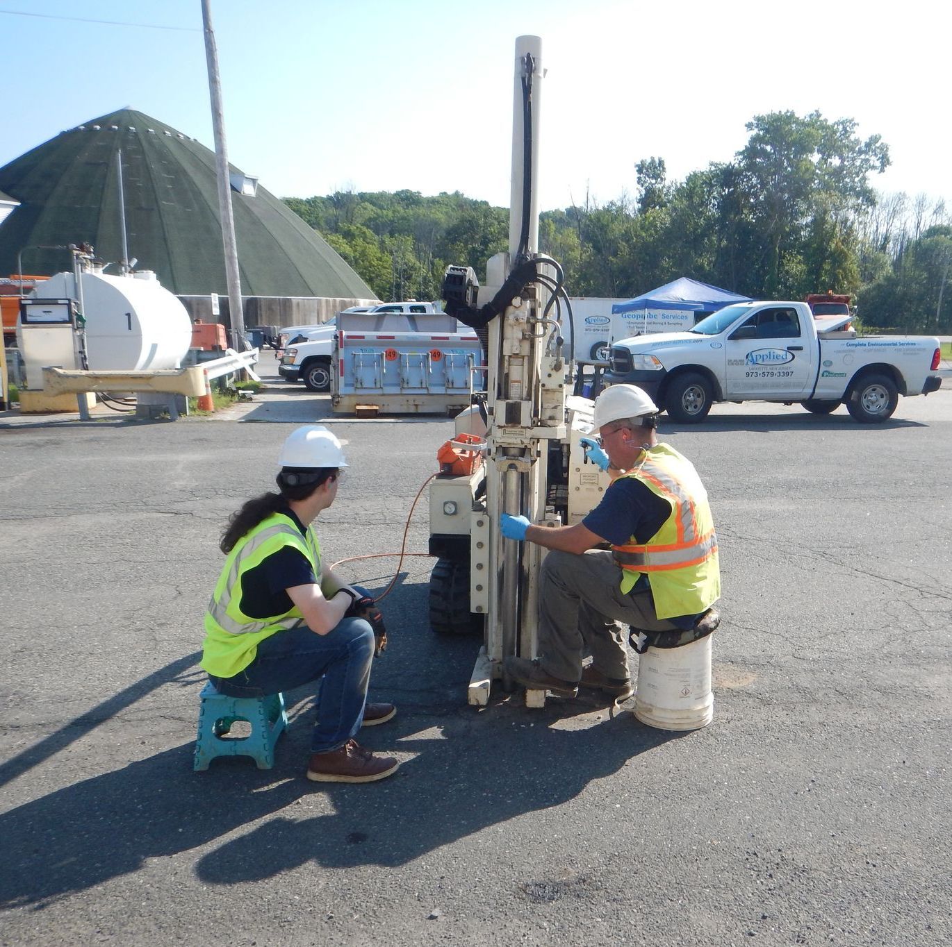 Two workers in safety gear operate a drilling machine on asphalt, likely for soil sampling.