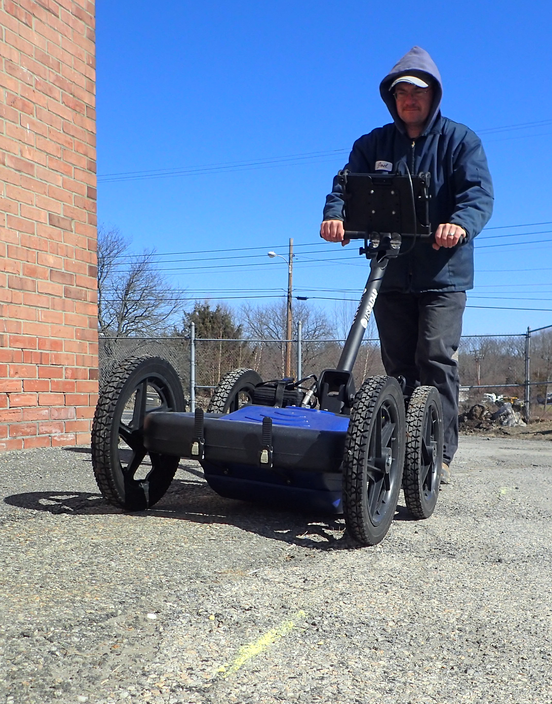 Man operating ground-penetrating radar on gravel next to a brick building.