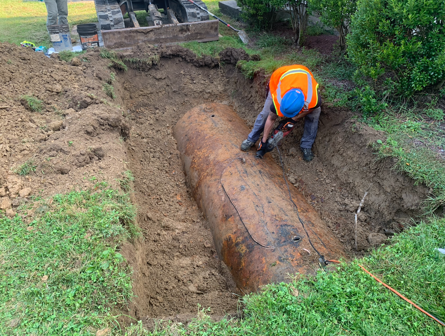 Man in orange vest cuts into buried metal tank in a muddy pit. Green grass surrounds.