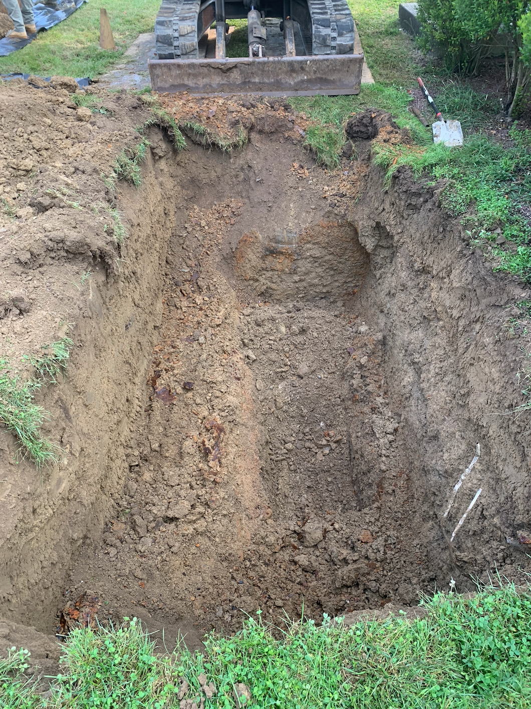Grave site, open pit dug in brown earth, stone headstone above. Green grass and vegetation at edges.