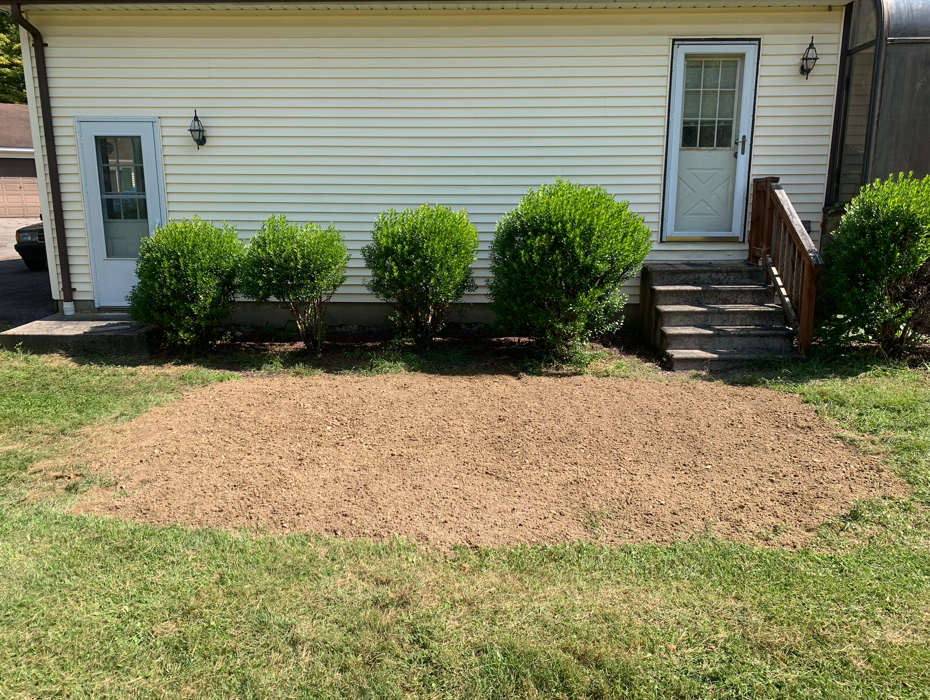 A sunny backyard shows a bed of mulch in front of a cream-colored house with green bushes and a staircase.