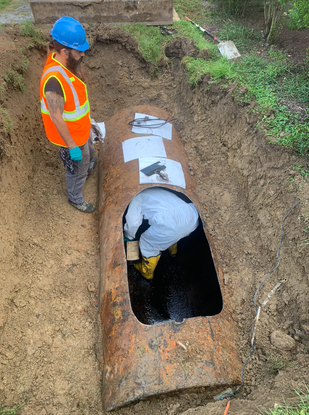 Two workers examining a rusted underground storage tank in a muddy excavation, one inside and one observing.