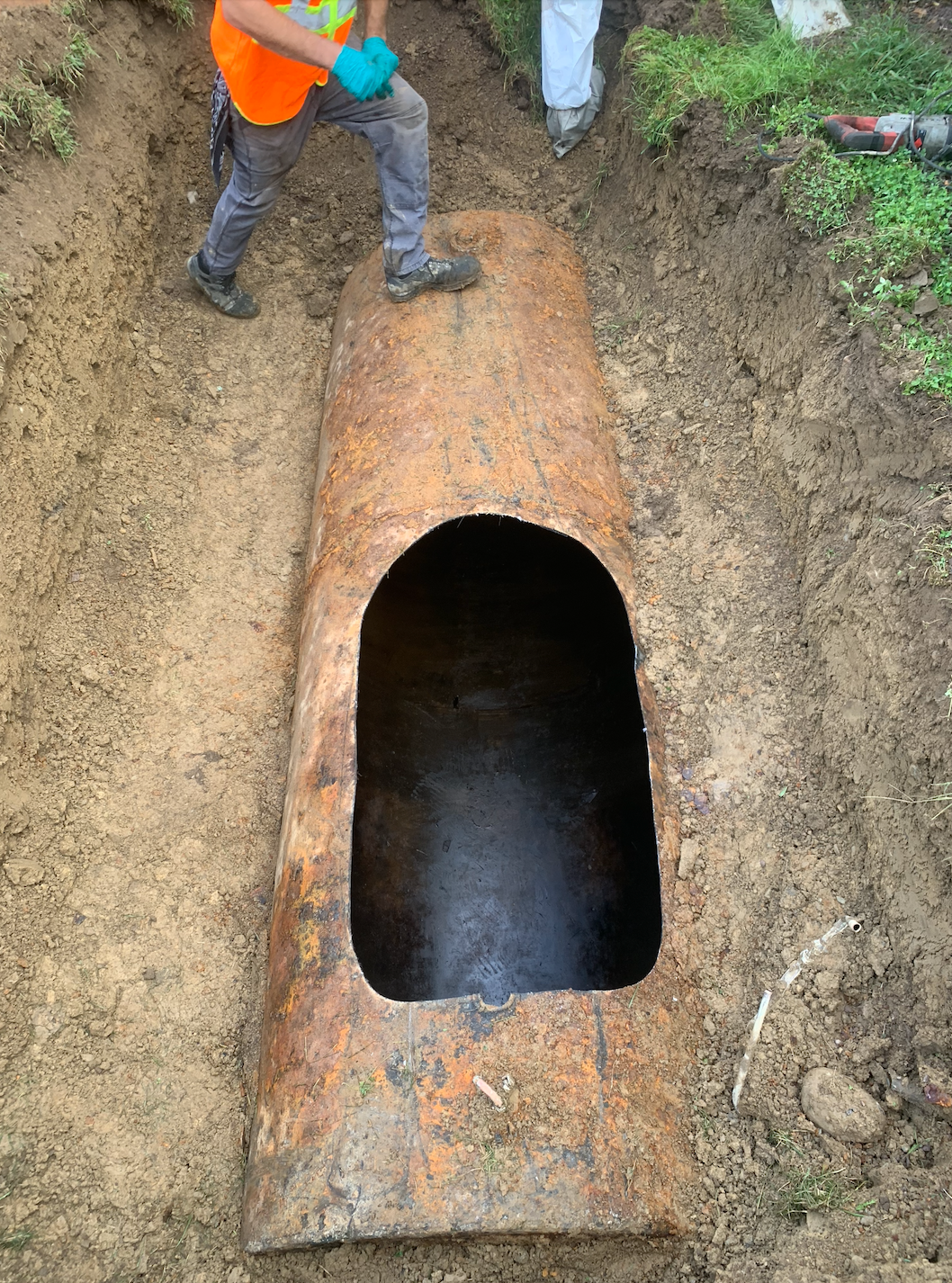 Workers inspecting a rusted metal tank in an excavation; orange safety vest visible.