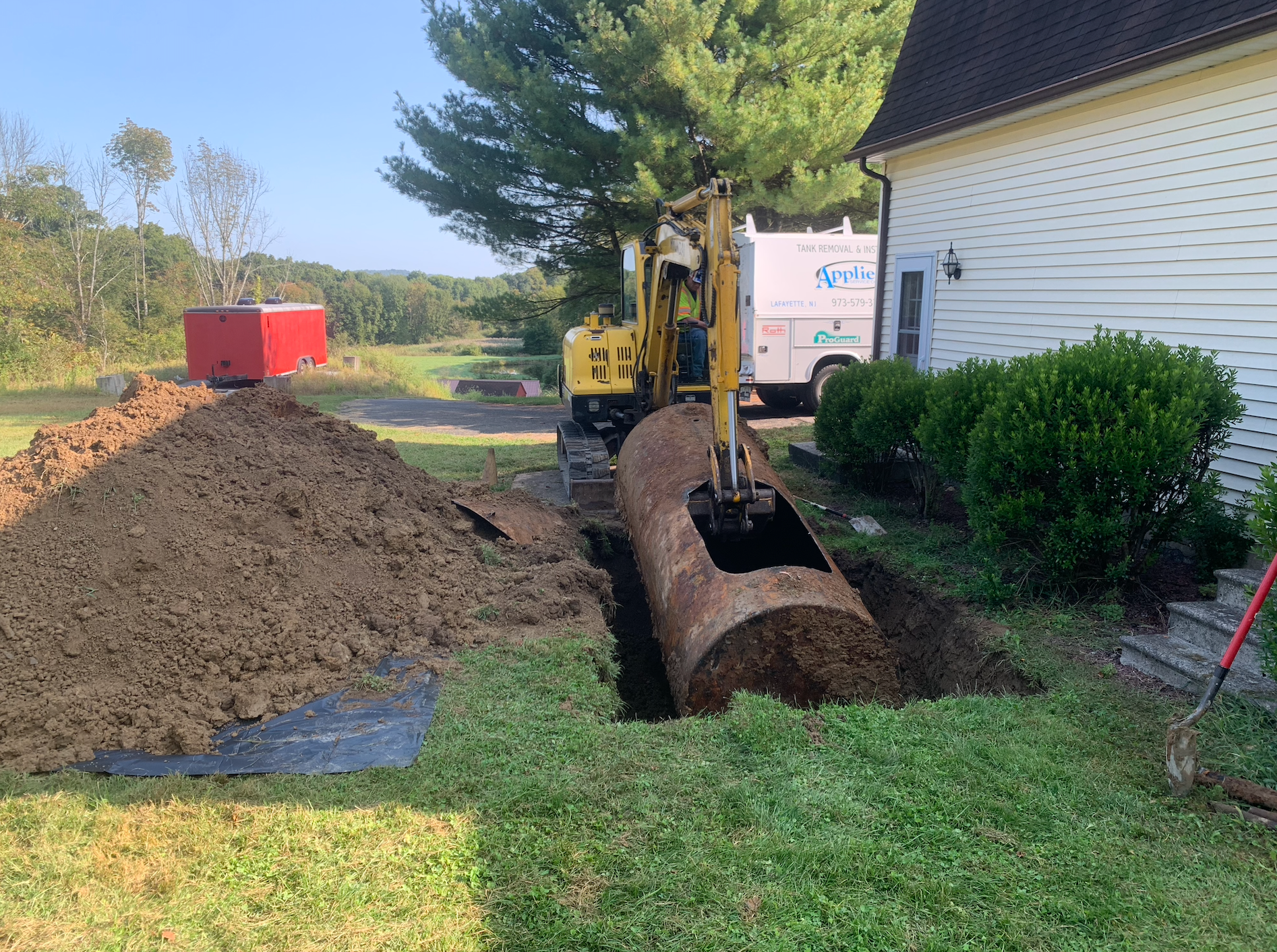 A small excavator removing an old, rusty fuel tank from a trench in a residential yard.